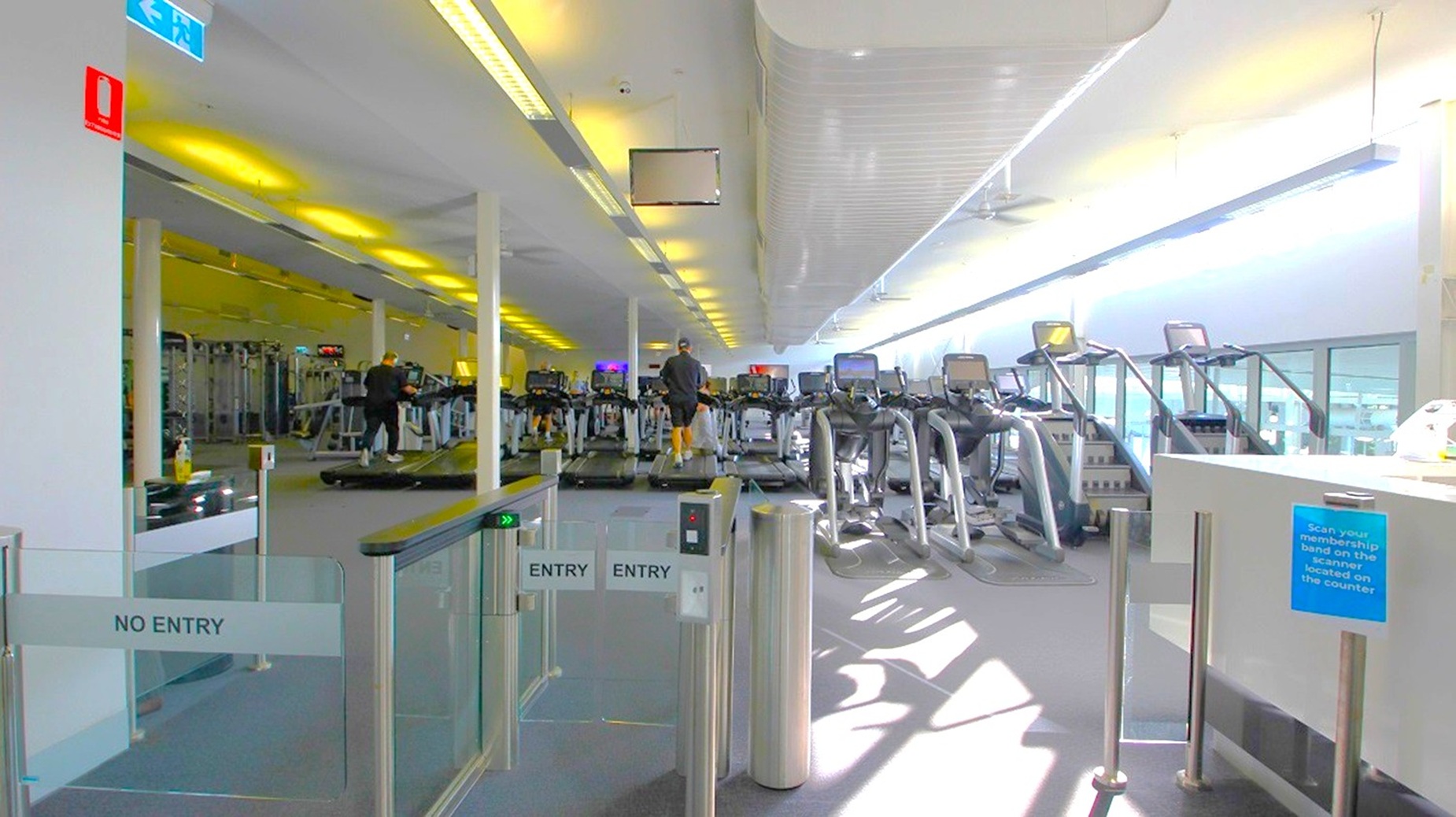 A gym interior with a row of treadmills on the right side, each equipped with digital screens. A person is using one of the treadmills. On the left, there are various gym machines and equipment. The entrance area in the foreground has gates marked "ENTRY" and "NO ENTRY." The ceiling is lined with fluorescent lights, and large windows on the right allow natural light to illuminate the space. The floor is carpeted in a neutral colour. A gym interior with a row of treadmills on the right side, each equipped with digital screens. A person is using one of the treadmills. On the left, there are various gym machines and equipment. The entrance area in the foreground has gates marked "ENTRY" and "NO ENTRY." The ceiling is lined with fluorescent lights, and large windows on the right allow natural light to illuminate the space. The floor is carpeted in a neutral colour.