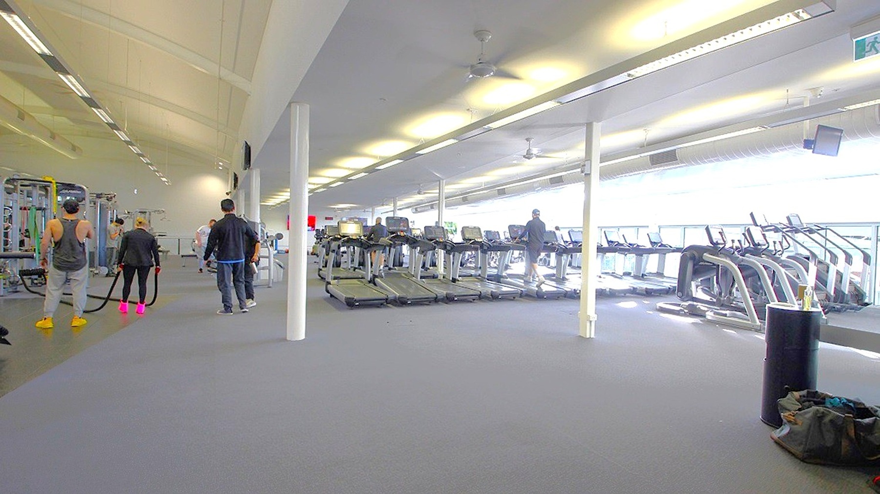 A spacious gym interior with a grey carpeted floor. On the left, several people are using exercise equipment, including a person holding battle ropes. In the centre, a row of treadmills is visible, with a few individuals using them. On the right, more cardio machines are lined up near large windows that let in natural light. The ceiling is high with fluorescent lights, and a white pillar is in the middle of the room. A black cylindrical object and a bag are on the floor in the foreground. A spacious gym interior with a grey carpeted floor. On the left, several people are using exercise equipment, including a person holding battle ropes. In the centre, a row of treadmills is visible, with a few individuals using them. On the right, more cardio machines are lined up near large windows that let in natural light. The ceiling is high with fluorescent lights, and a white pillar is in the middle of the room. A black cylindrical object and a bag are on the floor in the foreground.