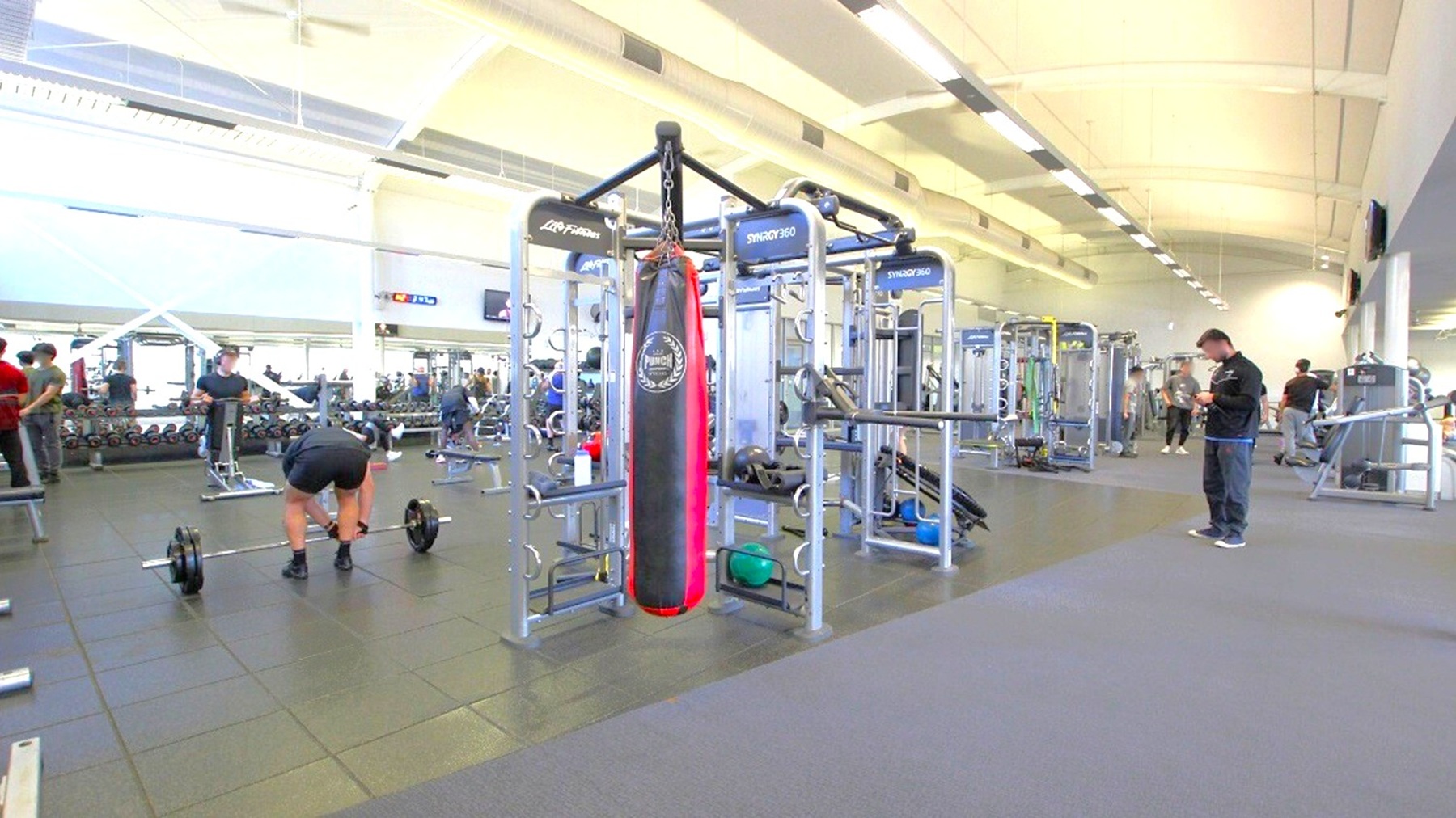 A spacious gym with various exercise equipment. In the centre, there's a large multi-functional machine with a red and black punching bag attached. To the left, a person is bending over a barbell, while others are using weights in the background. On the right, a person is standing, possibly checking a device. The floor is covered with dark rubber mats, and the ceiling is high with fluorescent lighting. The gym is well-lit with natural light coming through large windows. A spacious gym with various exercise equipment. In the centre, there's a large multi-functional machine with a red and black punching bag attached. To the left, a person is bending over a barbell, while others are using weights in the background. On the right, a person is standing, possibly checking a device. The floor is covered with dark rubber mats, and the ceiling is high with fluorescent lighting. The gym is well-lit with natural light coming through large windows.