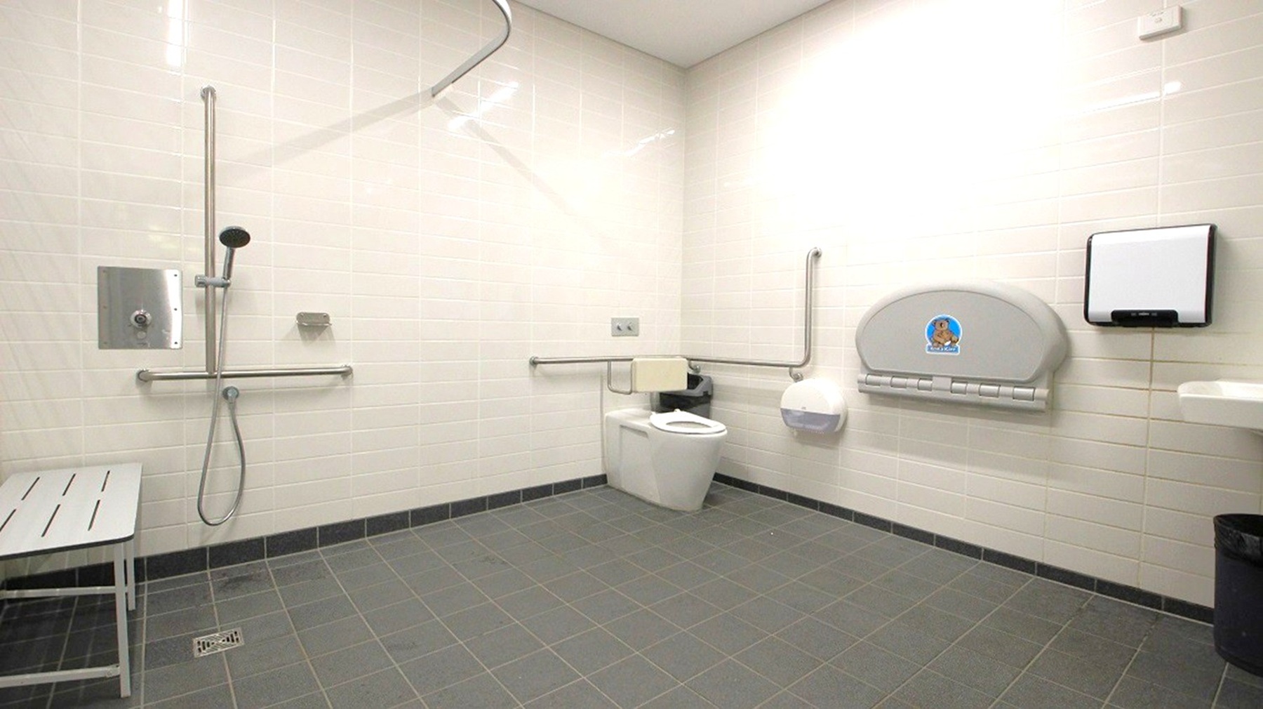 A spacious, accessible bathroom with light-coloured tiled walls and a grey tiled floor. On the left, there is a shower area with a handheld showerhead and a metal grab bar. A bench is positioned nearby. On the right, a toilet with support rails is visible, along with a baby changing station mounted on the wall. A paper towel dispenser and a waste bin are also present. The ceiling is plain and light-coloured. A spacious, accessible bathroom with light-coloured tiled walls and a grey tiled floor. On the left, there is a shower area with a handheld showerhead and a metal grab bar. A bench is positioned nearby. On the right, a toilet with support rails is visible, along with a baby changing station mounted on the wall. A paper towel dispenser and a waste bin are also present. The ceiling is plain and light-coloured.