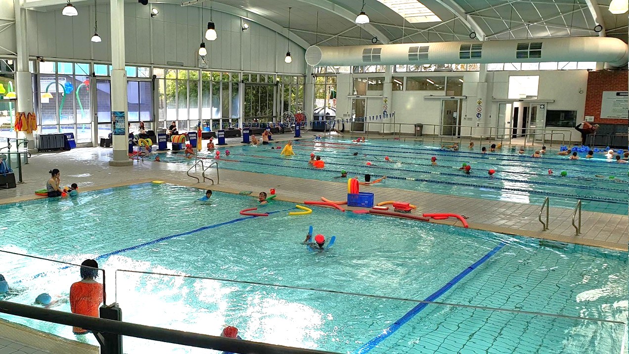 Indoor swimming pool with multiple lanes and people swimming. On the left, a smaller pool area with children and adults, some using floatation devices. The right side has a larger pool with more swimmers. The background features large windows allowing natural light, and a high, arched ceiling with hanging lights. Poolside, there are various swimming aids and equipment. The floor is tiled, and there are steps leading into the water. Indoor swimming pool with multiple lanes and people swimming. On the left, a smaller pool area with children and adults, some using floatation devices. The right side has a larger pool with more swimmers. The background features large windows allowing natural light, and a high, arched ceiling with hanging lights. Poolside, there are various swimming aids and equipment. The floor is tiled, and there are steps leading into the water.