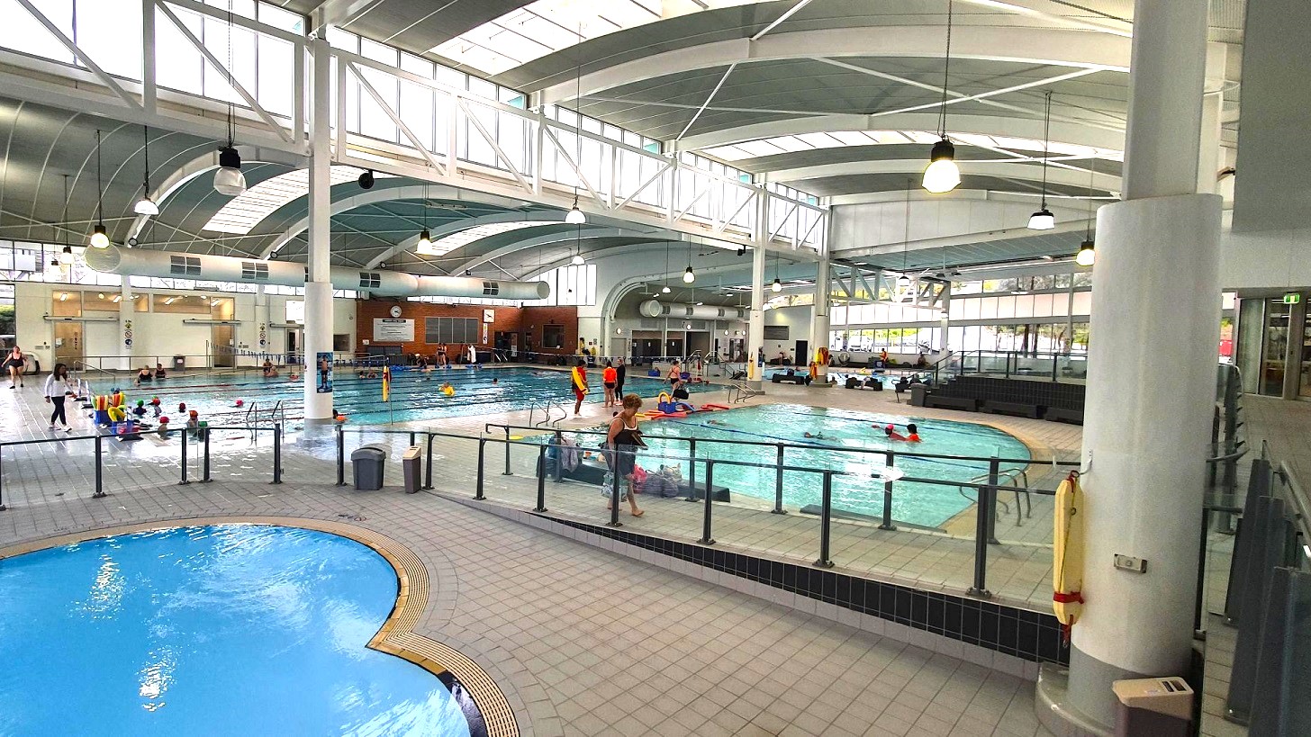 An indoor swimming pool complex with multiple pools. On the left, a smaller, round pool is visible with a tiled edge. In the centre, a larger rectangular pool is filled with people swimming and engaging in activities. The right side shows another pool area with people and a lifeguard. The ceiling is high with large windows and hanging lights, supported by white beams and columns. The floor is tiled, and there are railings separating the pool areas. An indoor swimming pool complex with multiple pools. On the left, a smaller, round pool is visible with a tiled edge. In the centre, a larger rectangular pool is filled with people swimming and engaging in activities. The right side shows another pool area with people and a lifeguard. The ceiling is high with large windows and hanging lights, supported by white beams and columns. The floor is tiled, and there are railings separating the pool areas.