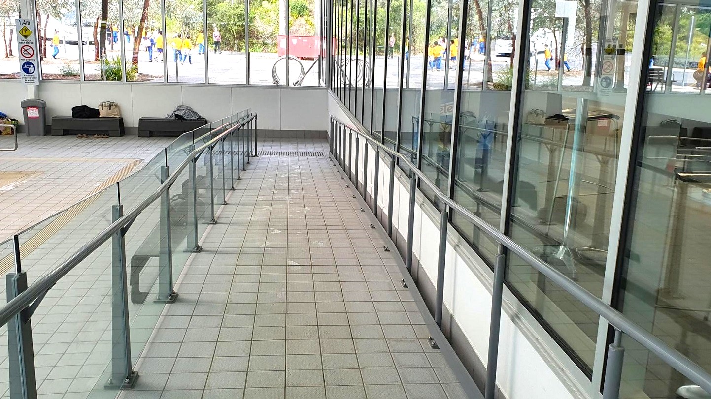 A tiled ramp with metal railings on both sides leads downwards. On the left, there are benches with bags placed on them and a rubbish bin nearby. Large windows on the right provide a view of people walking outside, wearing yellow shirts. The floor is tiled in a light grey and green pattern. Signs are visible on the wall to the left, and there are plants outside the windows. A tiled ramp with metal railings on both sides leads downwards. On the left, there are benches with bags placed on them and a rubbish bin nearby. Large windows on the right provide a view of people walking outside, wearing yellow shirts. The floor is tiled in a light grey and green pattern. Signs are visible on the wall to the left, and there are plants outside the windows.