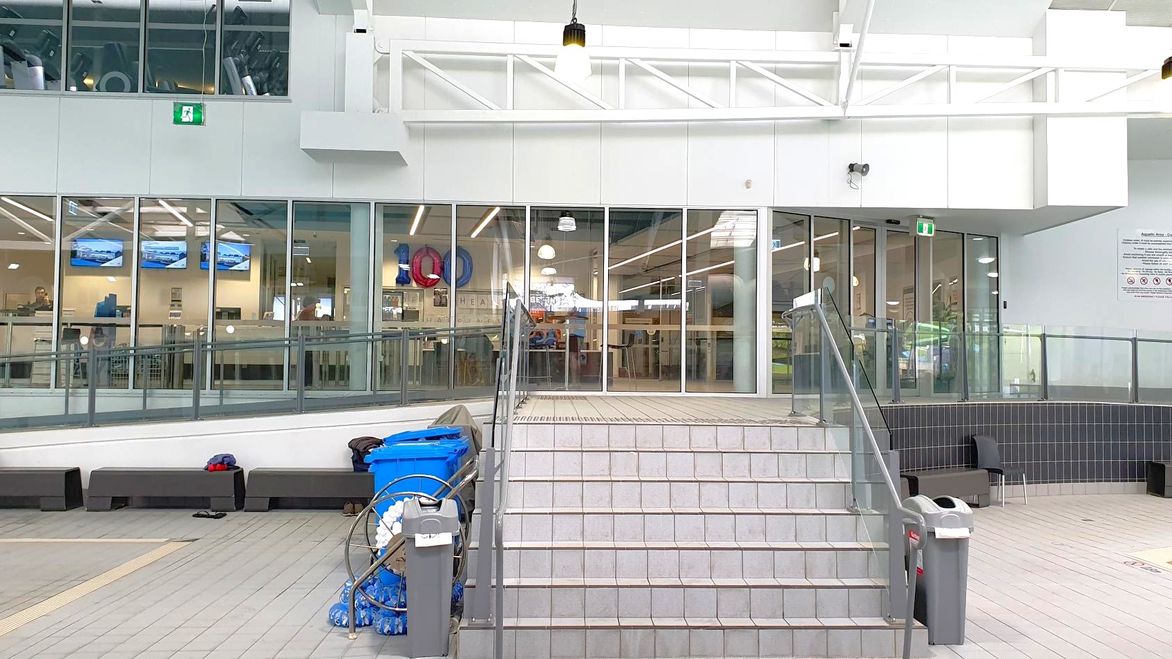 A set of grey tiled stairs leads up to a glass-walled interior space. The glass panels reveal a room with several screens displaying images and a few people inside. On the left, there are blue bins and cleaning equipment. The ceiling is white with exposed beams and hanging lights. The floor is tiled in grey, and there are black benches along the wall. A green exit sign is visible above the glass doors. A set of grey tiled stairs leads up to a glass-walled interior space. The glass panels reveal a room with several screens displaying images and a few people inside. On the left, there are blue bins and cleaning equipment. The ceiling is white with exposed beams and hanging lights. The floor is tiled in grey, and there are black benches along the wall. A green exit sign is visible above the glass doors.