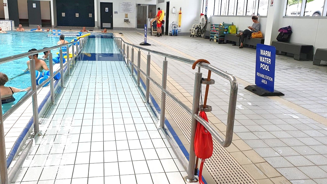 A swimming pool with a ramp on the left, featuring several people in the water using flotation devices. The right side has a tiled floor with a handrail and a red flotation device attached. A sign reads "Warm Water Pool Rehabilitation Area." In the background, a lifeguard stands near a wall, and a person sits on a bench reading. Large windows line the right wall, allowing natural light to enter. A swimming pool with a ramp on the left, featuring several people in the water using flotation devices. The right side has a tiled floor with a handrail and a red flotation device attached. A sign reads "Warm Water Pool Rehabilitation Area." In the background, a lifeguard stands near a wall, and a person sits on a bench reading. Large windows line the right wall, allowing natural light to enter.