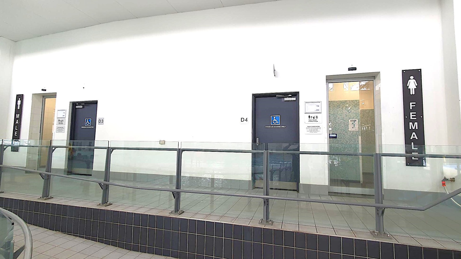 On the left, there is a door marked "MALE" with a sign above it. In the centre, there are two doors, D3 and D4, both with accessibility signs. On the right, a door marked "FEMALE" with a sign above it. A railing runs along the front, and the floor is tiled. The walls are plain white, and the ceiling is not visible. On the left, there is a door marked "MALE" with a sign above it. In the centre, there are two doors, D3 and D4, both with accessibility signs. On the right, a door marked "FEMALE" with a sign above it. A railing runs along the front, and the floor is tiled. The walls are plain white, and the ceiling is not visible.