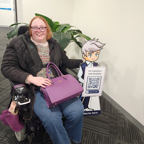 A woman in a wheelchair sits beside a Cérge cardboard cartoon cutout with a QR sign. A woman in a wheelchair sits beside a Cérge cardboard cartoon cutout with a QR sign.