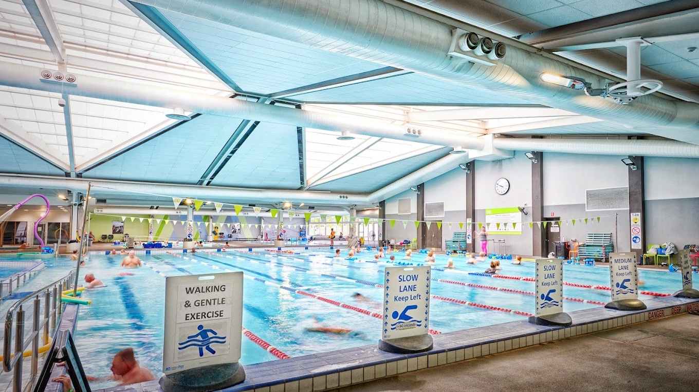 An indoor swimming pool is segmented into multiple lanes, demarcated by colourful floating ropes. Triangle banners stretch across the pool, and swimmers are visible in the water. On the left, a sign labelled 'WALKING & GENTLE EXERCISE' borders the raised pool deck, while the right side displays a 'SLOW LANE' sign. A ramp with handrails leading into the pool is positioned to the left. The ceiling is outfitted with large air conditioning vents, metal beams, and skylights.