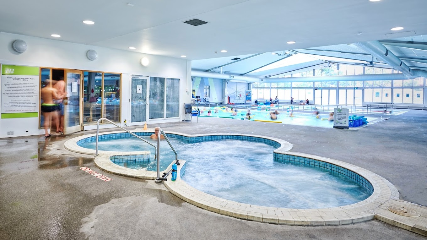 An indoor swimming facility features a spa in the foreground, equipped with steps and handrails, and a larger pool with a ramp and handrails in the background, occupied by swimmers. To the right, a large banner stands near a glass door, with poolside equipment scattered near the pool's edge. The ceiling is fitted with large air conditioning vents, metal beams, and skylights. A glass wall provides a view of the outside, and reminder posters are visible around the area.