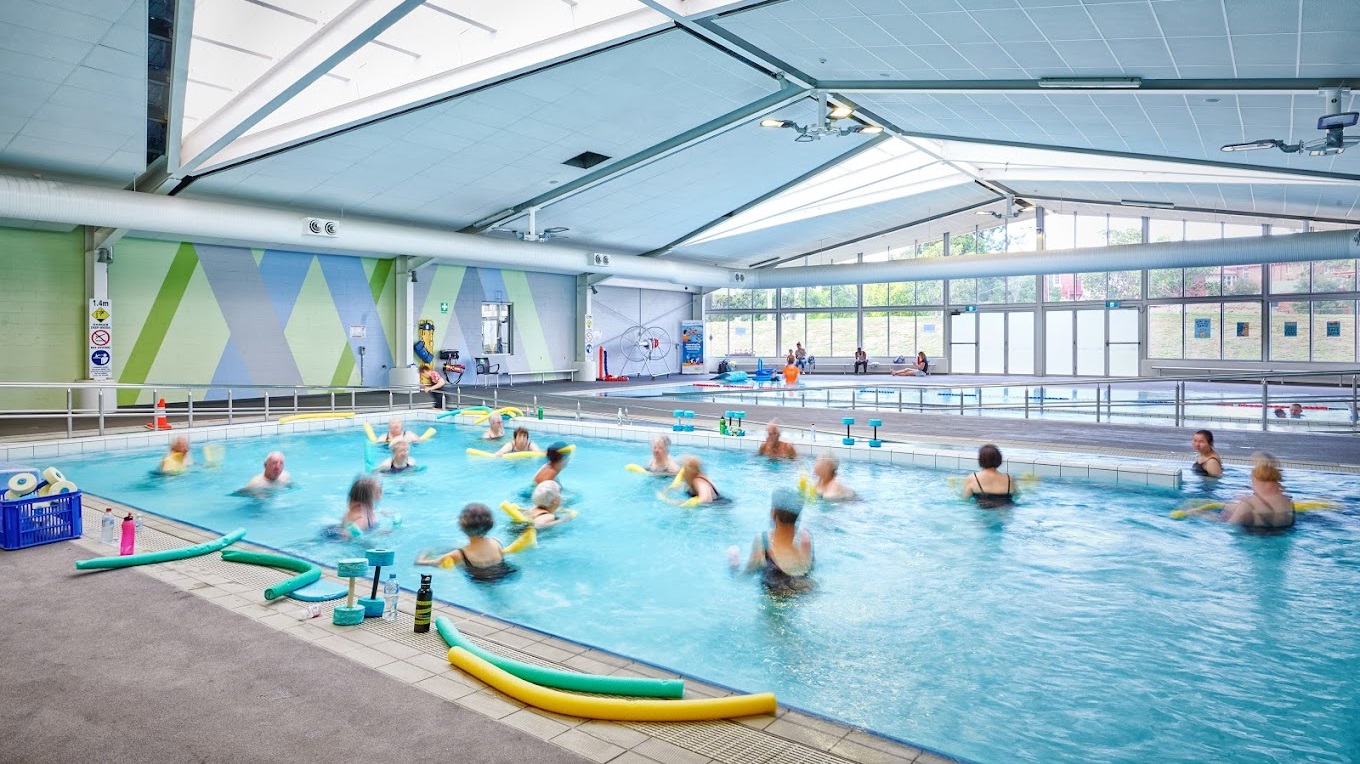 An indoor swimming pool, equipped with a ramp and handrails and currently occupied by swimmers, adjoins another pool sectioned by floating rope dividers. Noodle hoses, floaters, and other swimming accessories are scattered on the pool deck. The walls are painted green and blue with X-shaped patterns, while the ceiling features exposed metal beams, skylights, large air conditioning vents, and light fixtures. A glass wall at the far end encloses the aquatic facility.
