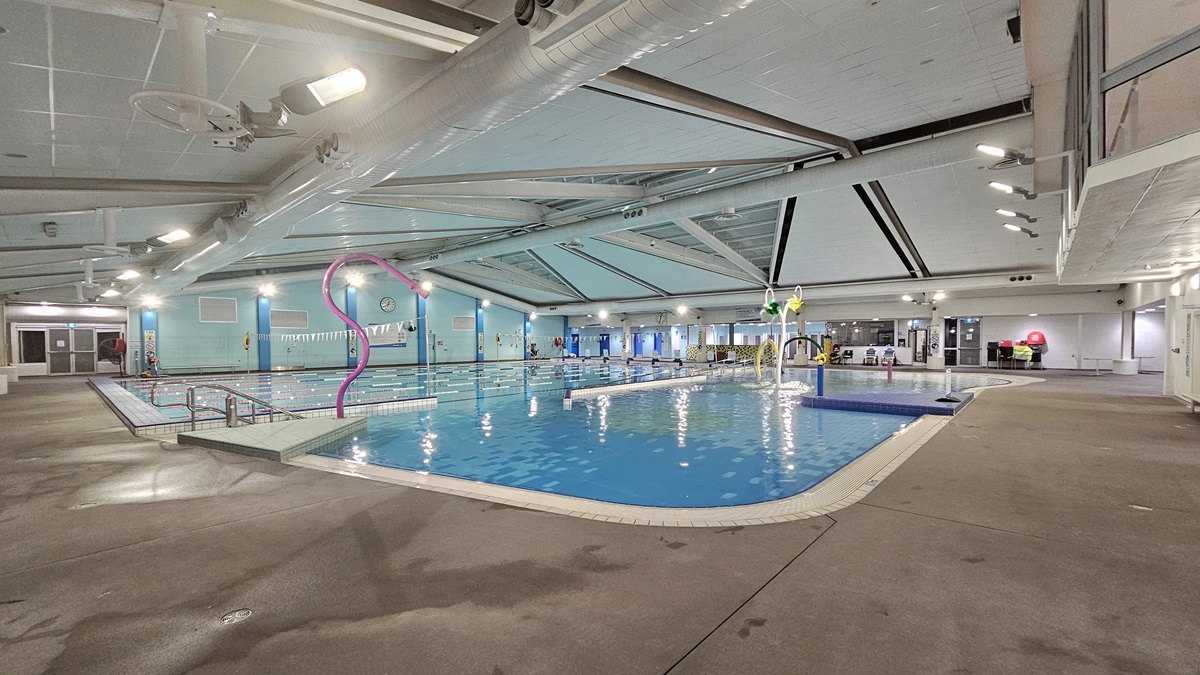 An indoor swimming facility features steps with handrails leading into a pool adorned with colourful hoops, sprayers, and a fountain. Adjacent to this is a larger pool bordered by floating ropes and triangular banners. The ceiling is equipped with large air conditioning vents, metal beams, and skylights. A glass wall provides views of the outside. Reminder posters are visible in the area. On the far right, chairs are stacked on the pool deck.
