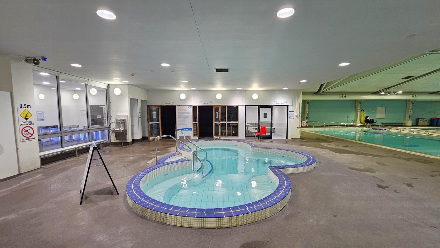 An indoor swimming facility includes a spa in the foreground with steps and handrails, and a larger pool equipped with a ramp and handrails. To the left of the spa, a freestanding banner and a bench chair are positioned against the glass wall. Behind the pool, three separate doors lead to additional facilities. The ceiling is outfitted with large air conditioning vents, metal beams, and skylights. Reminder posters are displayed around the area.