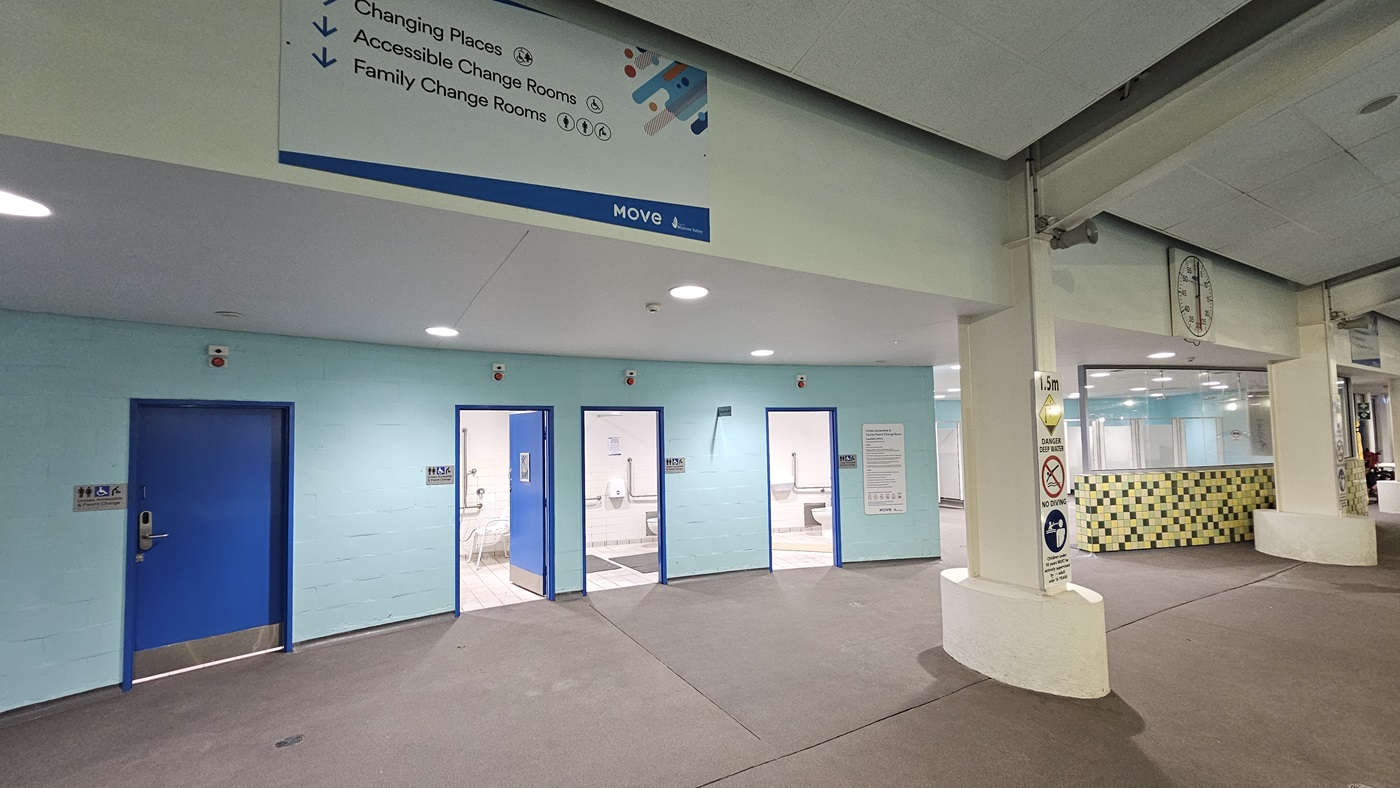 An indoor facility features a turquoise wall on the left with four blue doors, each labelled with gender access signs and signage for changing places and family rooms. On the right, there is another changing room with multiple cubicles, characterised by mosaic and glass walls. Large directional posters and a large round clock are affixed to the upper wall. Additional safety reminder posters are attached to the columns.
