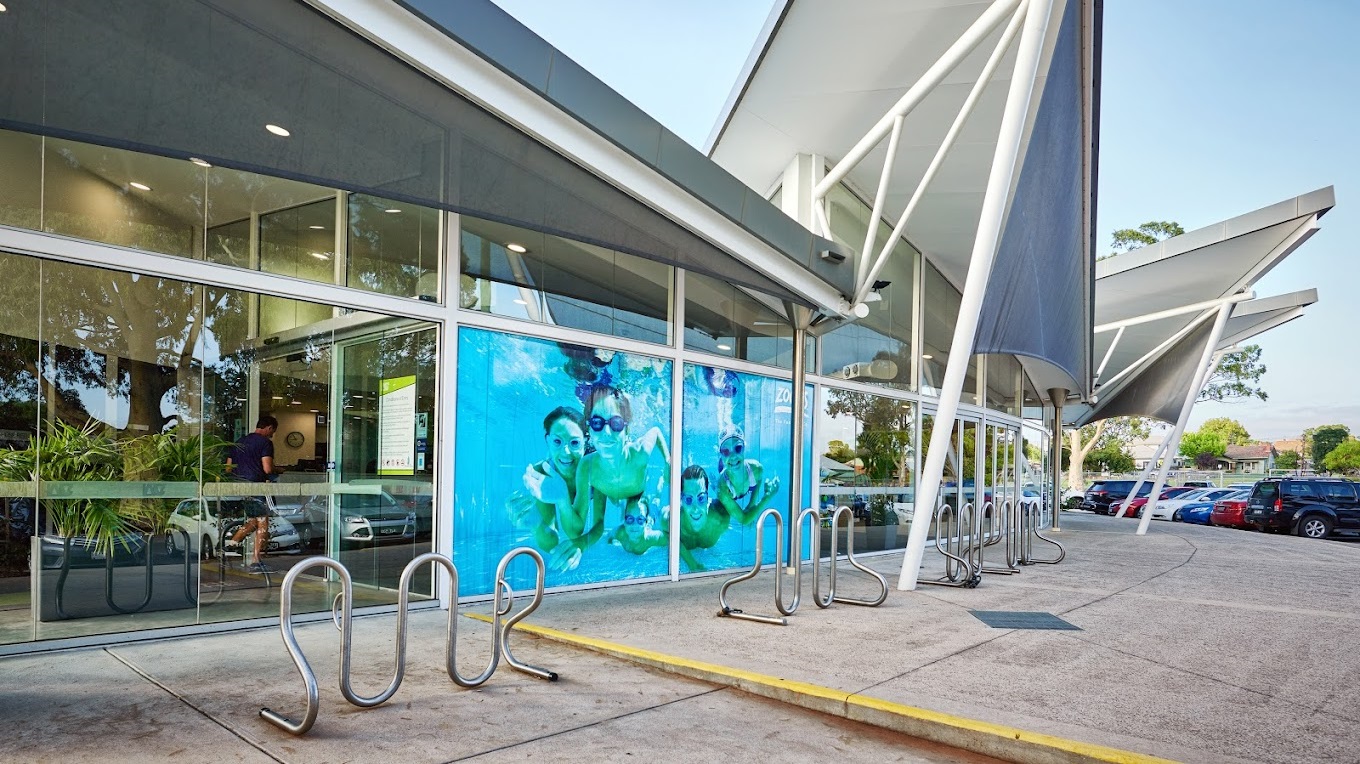 On the left, a modern building facade with a triangular shape roofing, reflective glass windows and a bold blue poster depicting swimmers. Silver bicycle racks curve outwards on the pavement. The right foreground shows a portion of a canopy structure with supporting poles, while parked cars line the background.