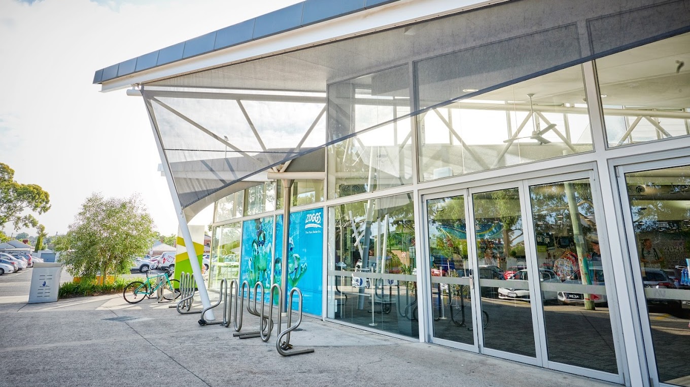 On the right, a modern building facade with a triangular shape roofing, reflective glass windows and a bold blue poster depicting swimmers. Silver bicycle racks curve outwards on the pavement. The right foreground shows a portion of a canopy structure with supporting poles, while parked cars line the background. Signage and trees are also visible.