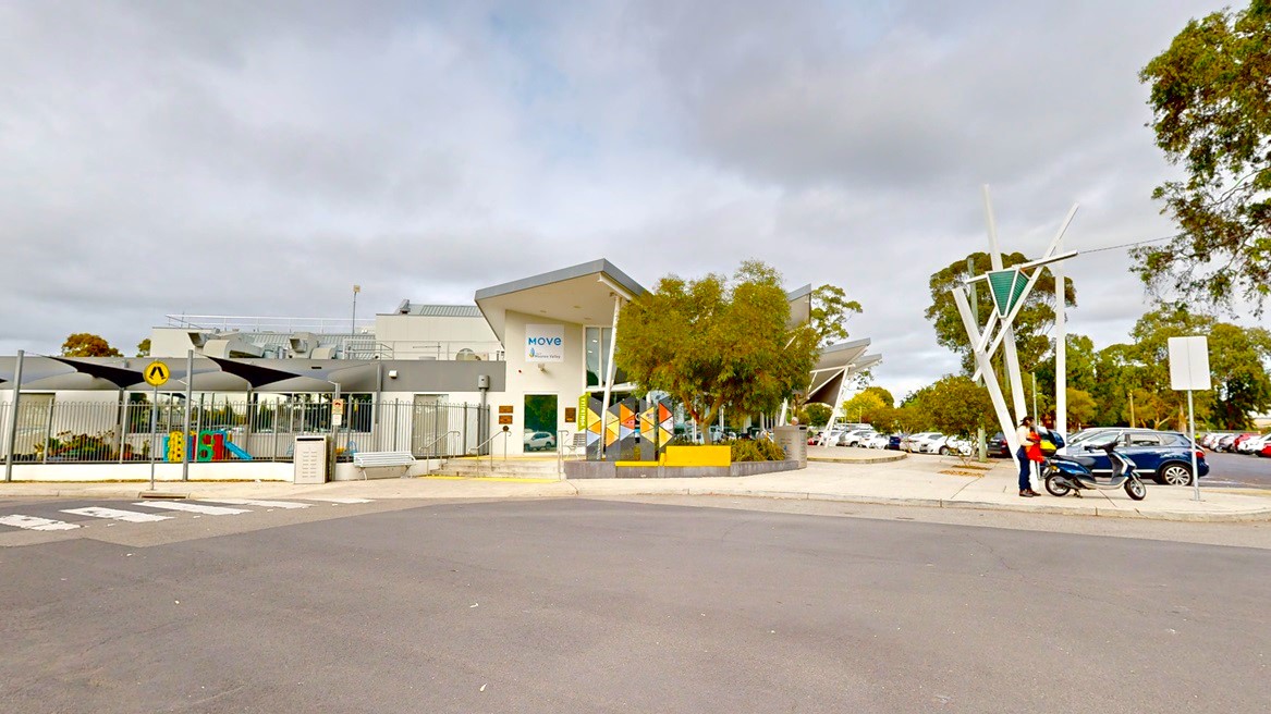 On the left, a low-set white building with pitched triangular roofing is enclosed by a white metal fence. Adjacent to the building are steps with handrails, and to the left, a planter box with trees and banners is situated. In the foreground, a road with a pedestrian lane marked in white is visible. Nearby, a car park hosts multiple vehicles of various colours. In front of the building, a bench chair, light post, mailbox, motorcycle, and pedestrians are also visible.