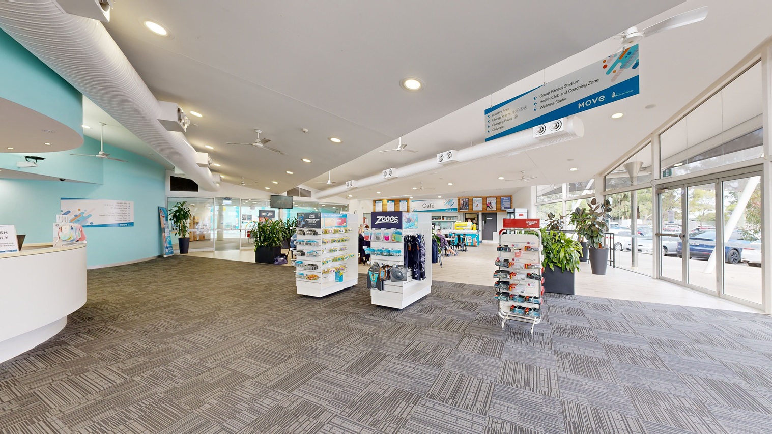 To the left, a reception desk with a white front features computers and brochures on top. Adjacent are rows of display racks showcasing goggles, swimwear, and accessories. Further to the left, a glass door provides a view of the car park outside. Indoor plants in large planter boxes are displayed around the area. The ceiling, adorned with light fixtures, posters, ceiling fans, and large air conditioning vents, is white, with light blue hues of the walls. The floor is covered with a grey carpet.