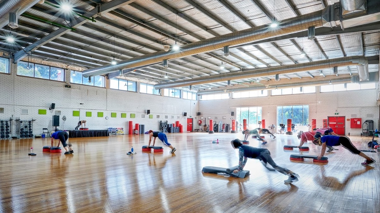A spacious indoor gym features polished timber flooring and a ceiling with exposed steel beams, large ceiling fans, silver insulation, and strip lighting. In the centre, individuals exercise on mats with step platforms. To the right, stationary bikes and red lockers line the wall. Equipment is scattered throughout the space. The wall is constructed from white bricks, and the central wall includes large glass windows.