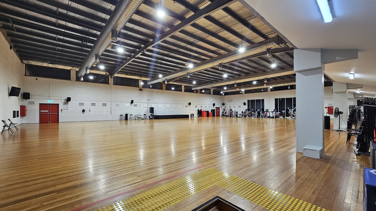 A spacious indoor gym features polished timber flooring and a ceiling decorated with exposed steel beams, large ceiling fans, silver insulation, and strip lighting. Chairs and various pieces of exercise equipment are arranged along the walls, which are made of white bricks. In the left corner, there is a two-tier set of steps fitted with truncated domes.