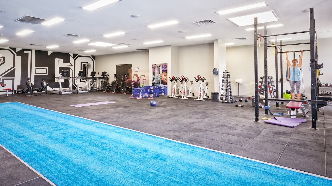 The interior of a gym is outfitted with equipment positioned on grey rubber flooring. To the left, weight machines and dumbbell racks are accompanied by smith machines. On the right, the area is equipped with spin machines, treadmills, and various cardio equipment. The background features mirrors and wall art, while a blue running track spans the foreground. The white ceiling above is fitted with recessed lighting.