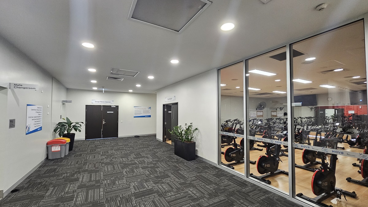 To the right is an enclosed space with timber floors and manual inward-opening doors, housing stationary bikes. To the left, white doors lead to male and female changing rooms. Rubbish bins and indoor plants are positioned against the wall. A black door is situated in the middle. The walls are white and adorned with posters. The ceiling is white, featuring air conditioning vents and recessed lighting, while the floor is covered with grey carpet.