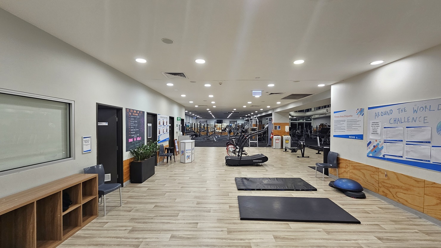 The interior of a modern gym features exercise equipment in the middle and at the far end. On the left, a wooden display rack and a chair are visible. Further to the left, three doors and a reception table are accompanied by notice boards and promotional banners. The foreground displays yoga mats and a balance ball. The flooring throughout is wood-patterned, combining timber and rubber mats. The ceiling is illuminated by recessed ceiling lights.