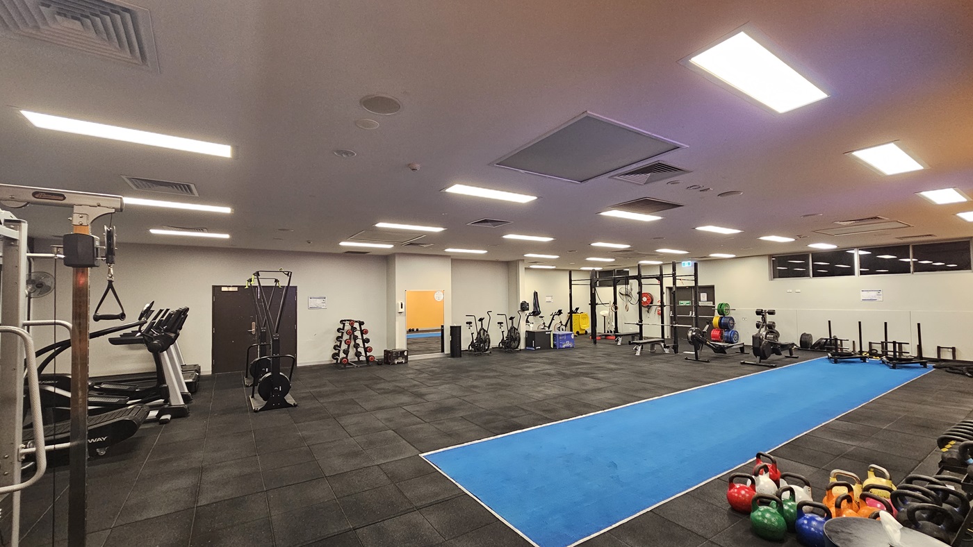 Fitness machines are arranged on the left, including treadmills, elliptical trainers, and Smith Machines. On the right, free weights, kettlebells, and a plyometric box are arranged. The central area features a long blue sprint mat. The ceiling is white with aircon vents, fluorescent lighting. The floor is covered with black rubber tiles, and mirrors are fitted on the far wall.