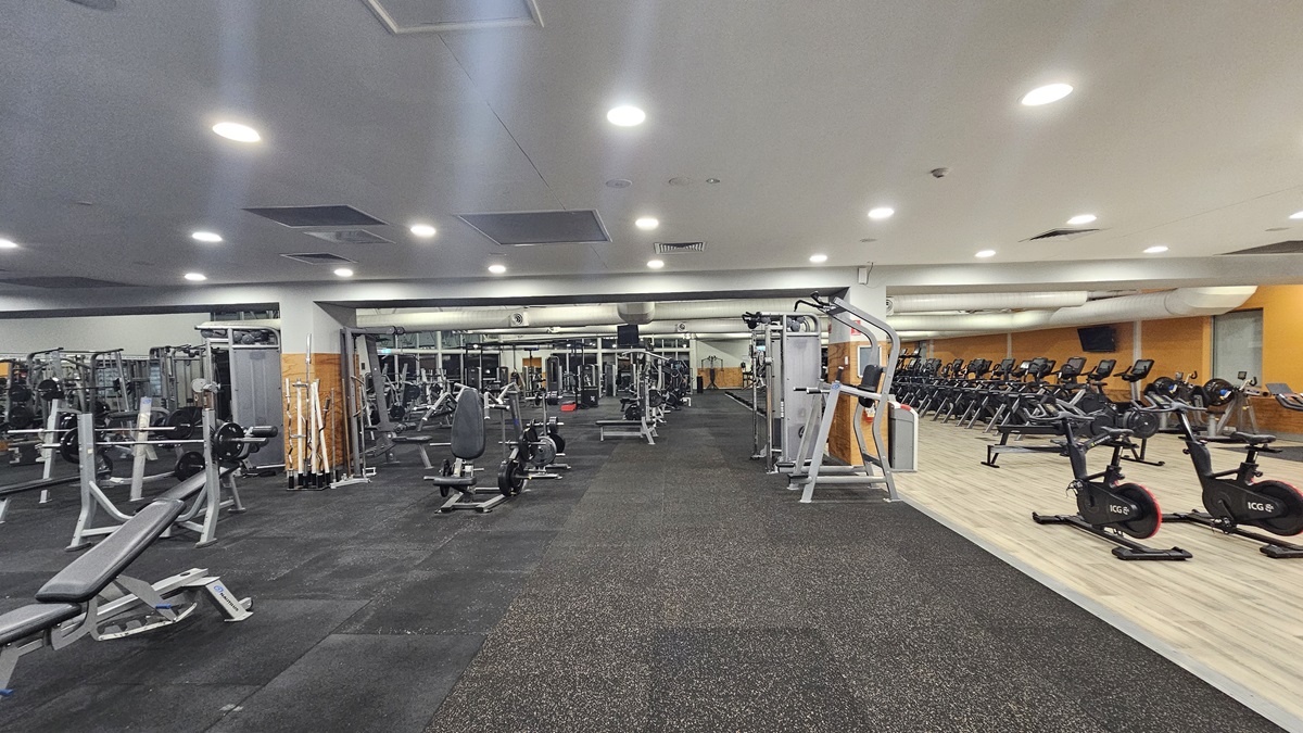 The interior of the gym features a variety of fitness equipment. To the left, there are weight benches, barbell racks, and sets of dumbbells. On the right, treadmills and stationary bikes are positioned. In the centre of the room, cable machines, lat pull-down machines, and bench machines are arranged. The entire floor is covered with grey padded flooring, and above, a white paneled ceiling is fitted with recessed lights.