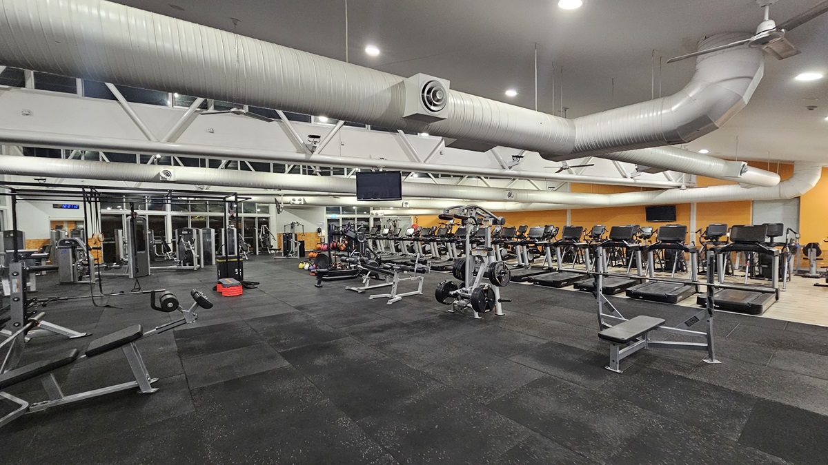 The interior of the well-equipped gym includes multiple workout stations on the left, featuring a Smith machine, a cable crossover machine, and several adjustable weight benches. Weight plates are neatly stacked on storage pegs attached to the machines. On the right, a row of treadmills faces the opposite direction, each equipped with a digital console. The floor is covered with black rubber tiles. The ceiling is painted white with ceiling fans, large aircon vents, and recessed lighting.