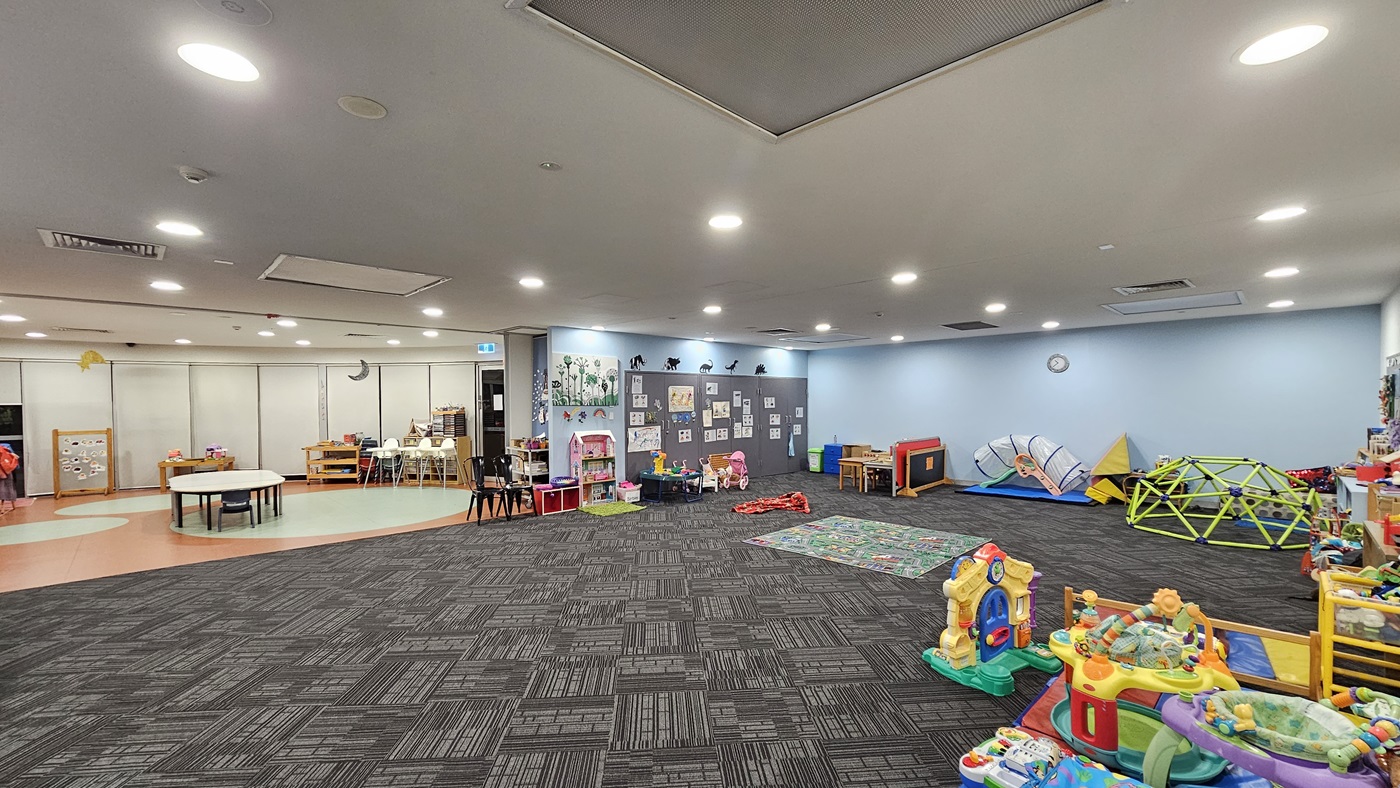 An interior view of a well-lit room features patterned grey carpet flooring. To the left, colourful children's artwork adorns the walls above shelving filled with books, alongside a play area with toys. To the right, there is a pram and additional toy storage. Shelves displaying toys are arranged against the walls. A colourful map carpet is positioned in the middle of the room. The ceiling is white, and equipped with recessed lighting and air conditioning vents.