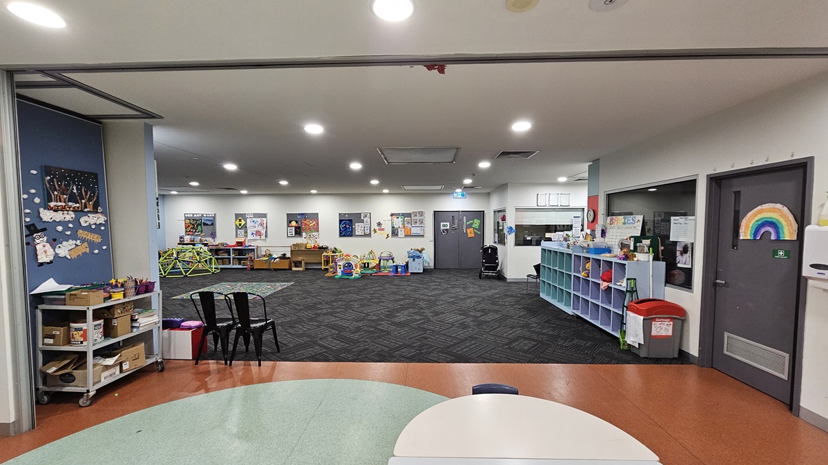 A spacious indoor area with a patterned grey carpet. To the left, a blue wall with children's artwork, shelving with toys, and a small table with chairs. On the right, a white wall and door with a rainbow motif. The foreground shows a white round table edge, while the ceiling is white with recessed lighting.