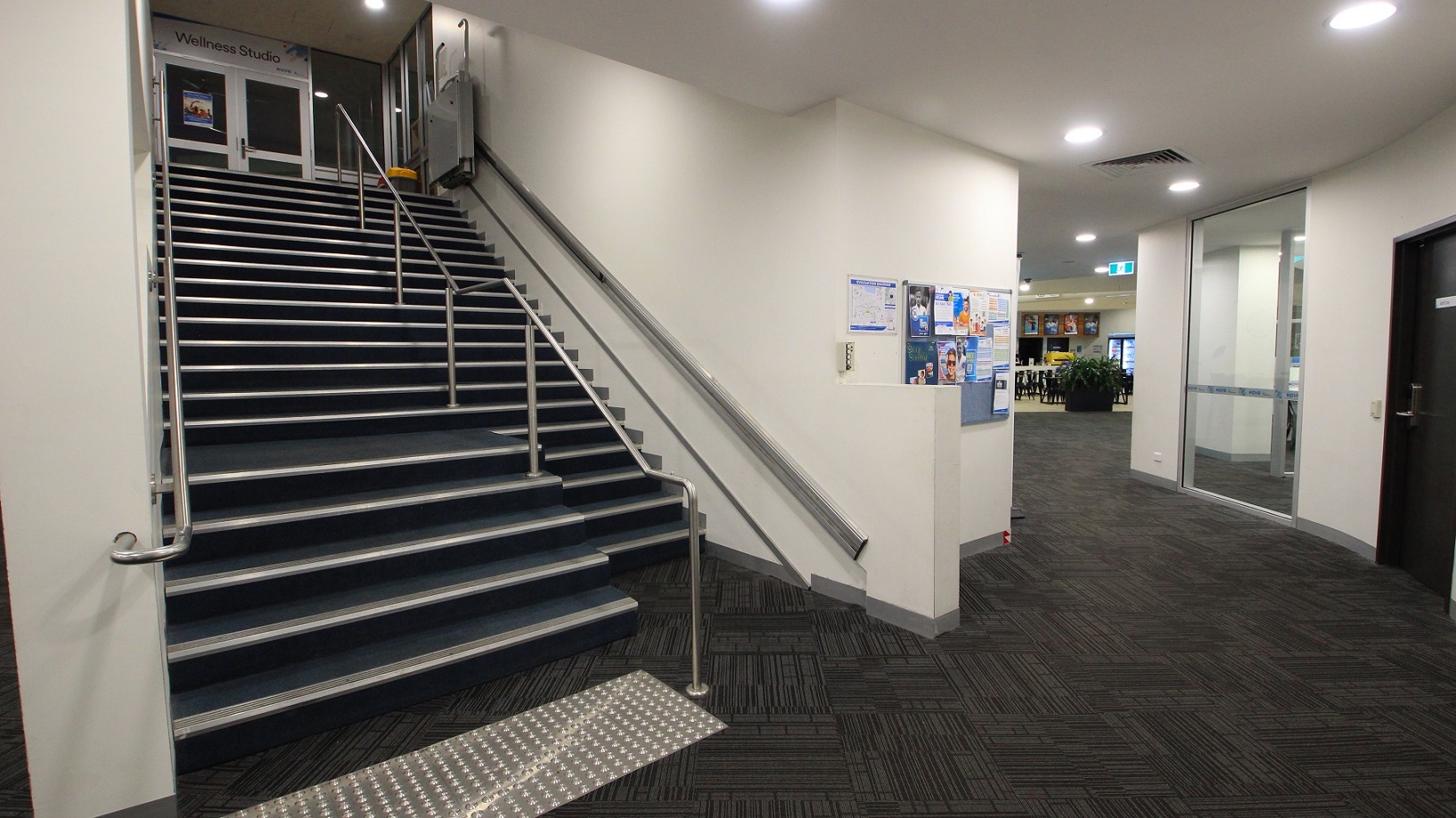 A corridor features a staircase on the left, equipped with handrails and truncated domes, leading to an upper level. A handrail runs parallel to the steps. On the right, a glass door opens into a room, next to another black door with a handle. The floor is patterned with dark geometric shapes, and the ceiling is fitted with recessed lighting. A noticeboard hangs on the back wall. In the background, a café is visible.
