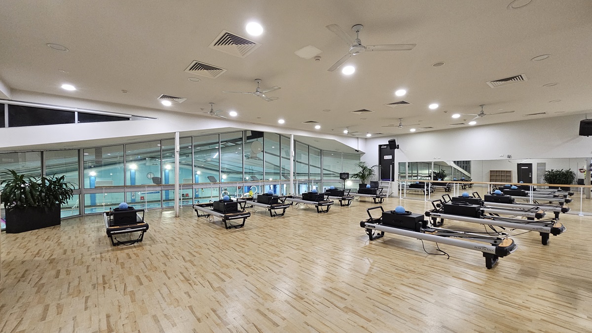 A spacious, well-lit room features a timber floor and a white ceiling equipped with multiple fans, recessed lights, and air conditioning. To the left, large windows offer a view of the aquatic facility below. To the right, a long mirror reflects the room's interior, which extends with visible equipment. Pilates reformer equipment is foregrounded, while indoor plants are positioned near the windows.