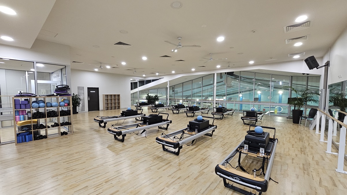 An indoor gym features Pilates reformers arranged centrally in rows. To the left, there is a rack holding various exercise balls and mats; to the right, large mirrors reflect the room. The white ceiling is equipped with recessed lights, ceiling fans, and air conditioning. The floor is polished timber. Another display rack is positioned near a black door further inside. The wall on the right offers a view of the pool below.