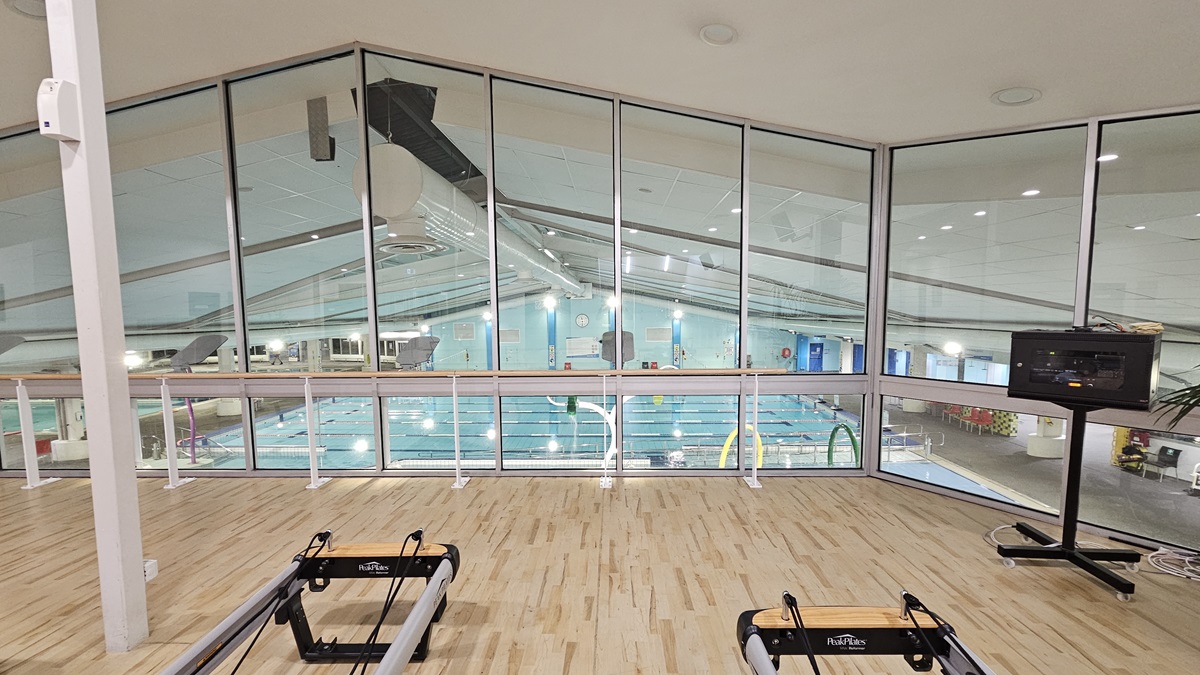 An indoor gym displays Pilates reformers arranged on the left, with a manual glass door entrance adjacent. To the left, there is a rack, a rubbish bin, and large indoor plants placed next to a chair. The white ceiling is fitted with recessed lights, ceiling fans, and air conditioning. The floor is polished timber. Another display rack is located near a black door further inside. The wall on the left features a mirror reflecting the interior.