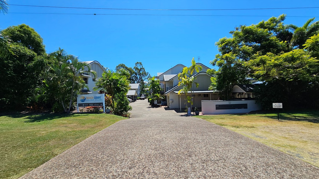 A paved driveway stretches from the foreground to the middle, flanked by green grass on both sides. To the left, a signboard for "Beach Court Holiday Villas" is set amidst dense palm trees. To the right, a white building with a dark sign is partially visible behind smaller trees. In the middle, modern two-storey buildings with light facades and peaked roofs line the driveway, with additional trees and greenery between them. A paved driveway stretches from the foreground to the middle, flanked by green grass on both sides. To the left, a signboard for "Beach Court Holiday Villas" is set amidst dense palm trees. To the right, a white building with a dark sign is partially visible behind smaller trees. In the middle, modern two-storey buildings with light facades and peaked roofs line the driveway, with additional trees and greenery between them.