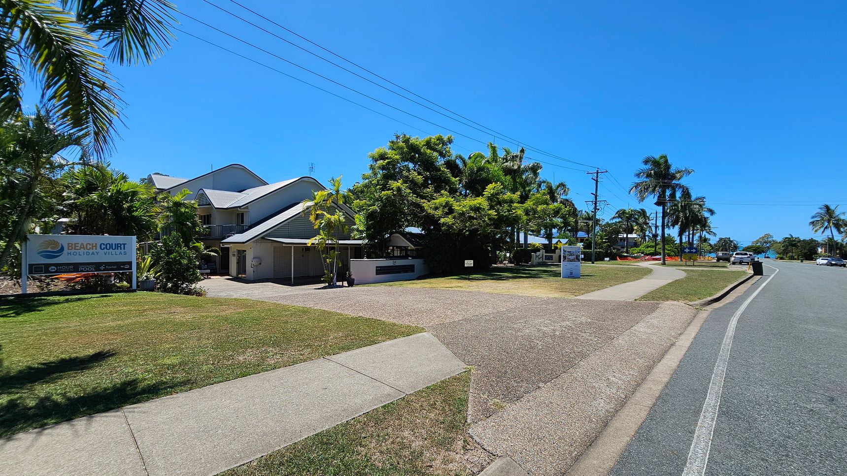 On the left, there is a two-storey building with a grey roof surrounded by palm trees and greenery. A signboard reading “Beach Court Holiday Villas” stands on the grass. On the right, there is a paved road bordered by more palm trees, parked cars, and a footpath. In the middle, a driveway on a pebbled surface leads to the building. The background features more trees and low-lying structures. The foreground contains grassy patches and concrete footpaths. On the left, there is a two-storey building with a grey roof surrounded by palm trees and greenery. A signboard reading “Beach Court Holiday Villas” stands on the grass. On the right, there is a paved road bordered by more palm trees, parked cars, and a footpath. In the middle, a driveway on a pebbled surface leads to the building. The background features more trees and low-lying structures. The foreground contains grassy patches and concrete footpaths.