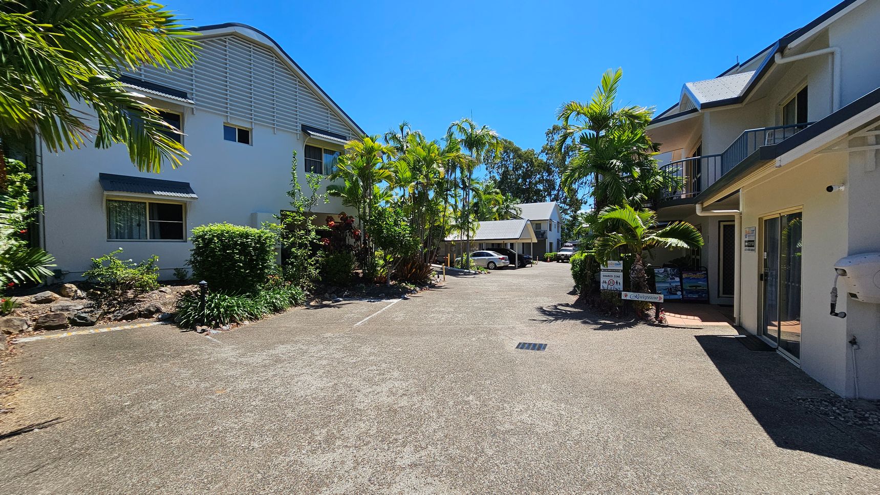 On the left, there is a white two-storey building with small awnings over the windows and greenery including bushes and palm trees. On the right, there is a similar building with balconies, glass doors, and potted plants near the entrance. In the middle, a paved area extends towards a smaller building and parked cars. The background includes more trees and shrubs. The foreground features a concrete surface with a drainage grate. On the left, there is a white two-storey building with small awnings over the windows and greenery including bushes and palm trees. On the right, there is a similar building with balconies, glass doors, and potted plants near the entrance. In the middle, a paved area extends towards a smaller building and parked cars. The background includes more trees and shrubs. The foreground features a concrete surface with a drainage grate.