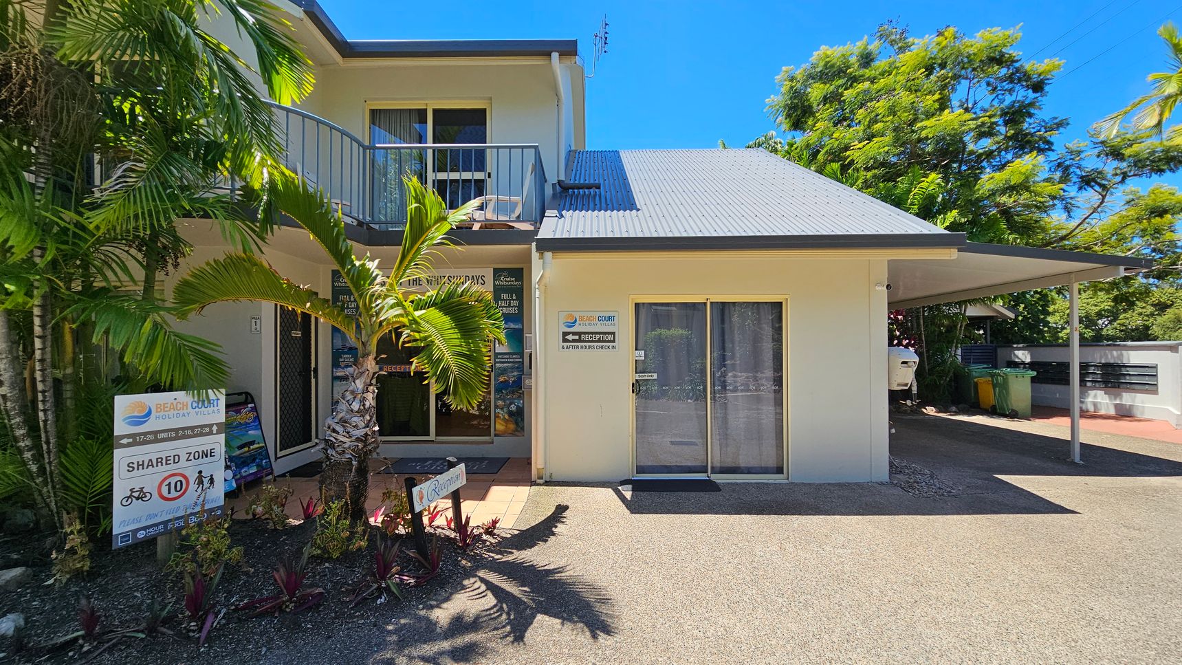On the left, there is a two-storey cream-coloured building with a balcony, sliding glass doors, and a small palm tree in front. Below, a sign displays “Beach Court Holiday Villas” and shared zone instructions near garden beds with small plants. On the right, a single-storey section features a sliding glass door with a curtain and a covered carport. The foreground consists of a textured, pebbled surface, while the background includes trees and greenery. On the left, there is a two-storey cream-coloured building with a balcony, sliding glass doors, and a small palm tree in front. Below, a sign displays “Beach Court Holiday Villas” and shared zone instructions near garden beds with small plants. On the right, a single-storey section features a sliding glass door with a curtain and a covered carport. The foreground consists of a textured, pebbled surface, while the background includes trees and greenery.