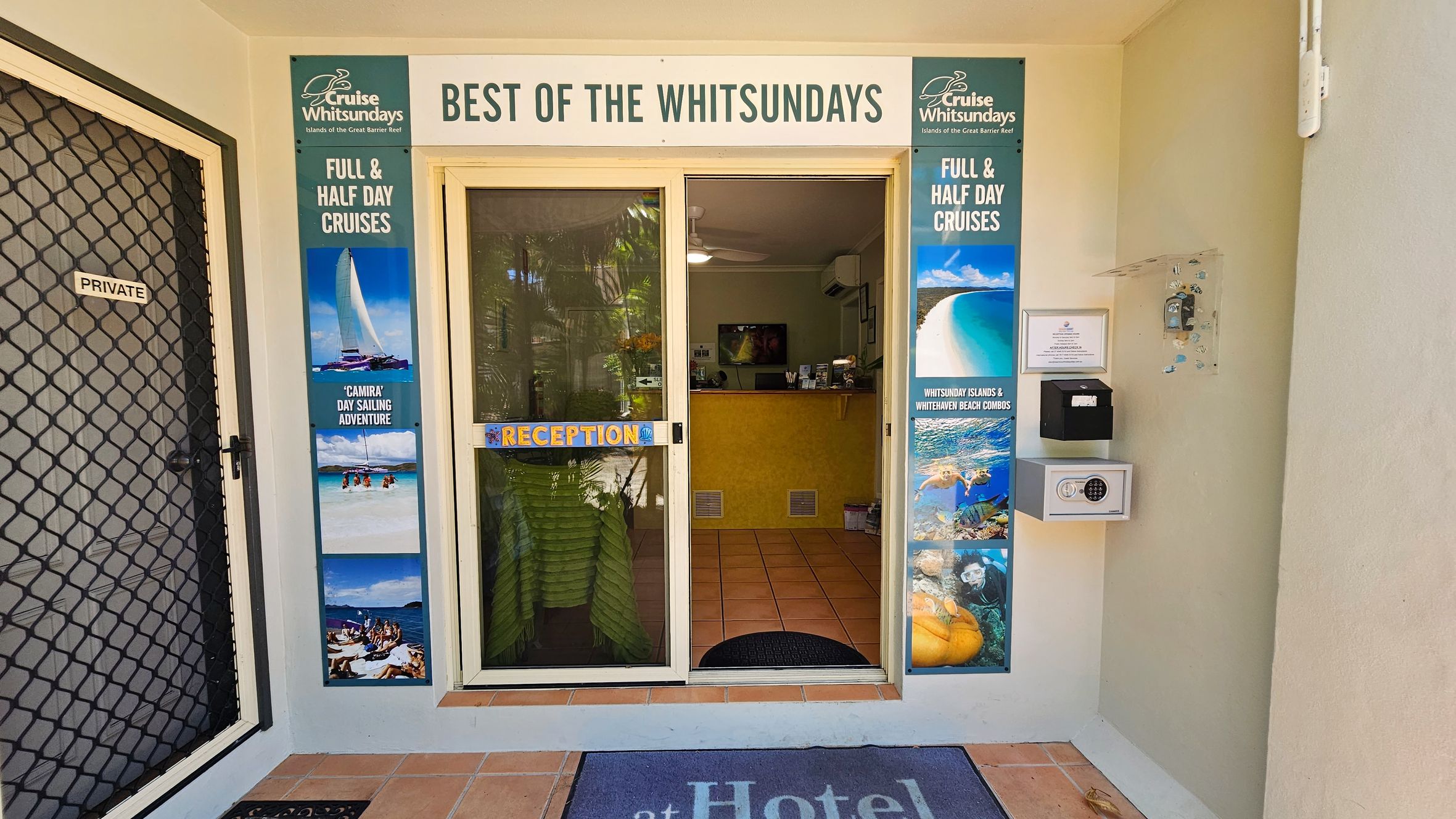 On the left, there is a black mesh door labelled "Private" beside a vertical poster advertising cruises, with images of sailing and beaches. On the right, a wall-mounted safe, noticeboard, and another vertical cruise poster showing beach scenes and marine life are visible. In the middle, glass sliding doors with a "Reception" sign lead to a tiled room with a yellow desk, and ceiling fans. The floor is terracotta tiles, with a dark mat at the entrance. The ceiling inside is white and flat. On the left, there is a black mesh door labelled "Private" beside a vertical poster advertising cruises, with images of sailing and beaches. On the right, a wall-mounted safe, noticeboard, and another vertical cruise poster showing beach scenes and marine life are visible. In the middle, glass sliding doors with a "Reception" sign lead to a tiled room with a yellow desk, and ceiling fans. The floor is terracotta tiles, with a dark mat at the entrance. The ceiling inside is white and flat.