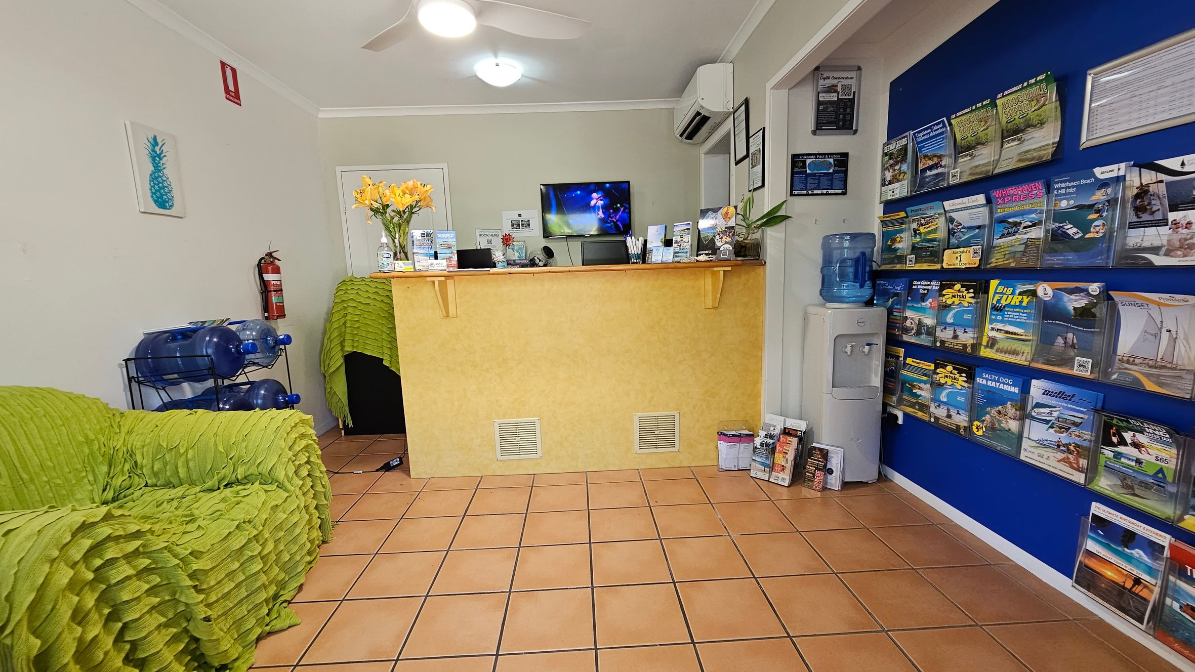 On the left, a lime-green textured sofa sits near a metal rack holding large blue water bottles. In the middle, there is a yellow reception desk with brochures, a vase of yellow flowers, and a wall-mounted television above it. On the right, a bright blue wall displays brochures in organised rows, alongside a water cooler. The floor is tiled in terracotta-style squares, and the ceiling is white with recessed lighting and a ceiling fan. On the left, a lime-green textured sofa sits near a metal rack holding large blue water bottles. In the middle, there is a yellow reception desk with brochures, a vase of yellow flowers, and a wall-mounted television above it. On the right, a bright blue wall displays brochures in organised rows, alongside a water cooler. The floor is tiled in terracotta-style squares, and the ceiling is white with recessed lighting and a ceiling fan.