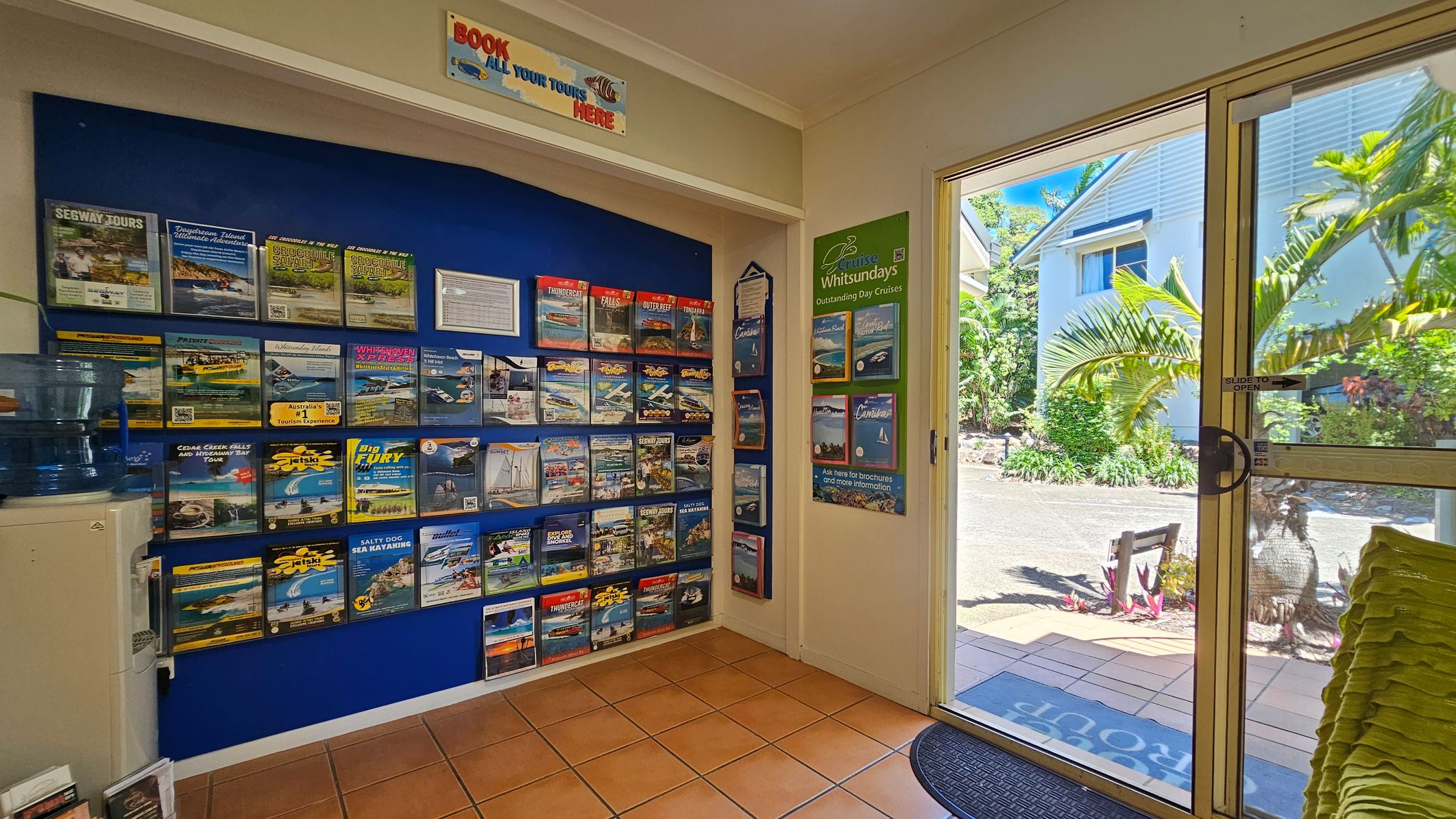 On the left, a blue wall is covered with neatly arranged brochures advertising tours and activities, with a water cooler positioned beneath them. In the middle, a sign above reads "Book all your tours here" in bold text. On the right, glass sliding doors open to an outdoor area with tropical plants, a paved path, and a white building visible in the background. The floor is tiled with terracotta-coloured squares, and the ceiling is white and flat. On the left, a blue wall is covered with neatly arranged brochures advertising tours and activities, with a water cooler positioned beneath them. In the middle, a sign above reads "Book all your tours here" in bold text. On the right, glass sliding doors open to an outdoor area with tropical plants, a paved path, and a white building visible in the background. The floor is tiled with terracotta-coloured squares, and the ceiling is white and flat.