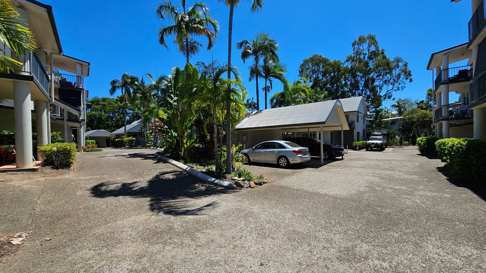 A residential area with two-storey buildings on the left and right, featuring balconies and supported by columns. In the centre, a driveway leads to a carport with a silver car parked underneath. Lush greenery, including palm trees and shrubs, surrounds the area. The ground is paved, and the sky is clear. A residential area with two-storey buildings on the left and right, featuring balconies and supported by columns. In the centre, a driveway leads to a carport with a silver car parked underneath. Lush greenery, including palm trees and shrubs, surrounds the area. The ground is paved, and the sky is clear.