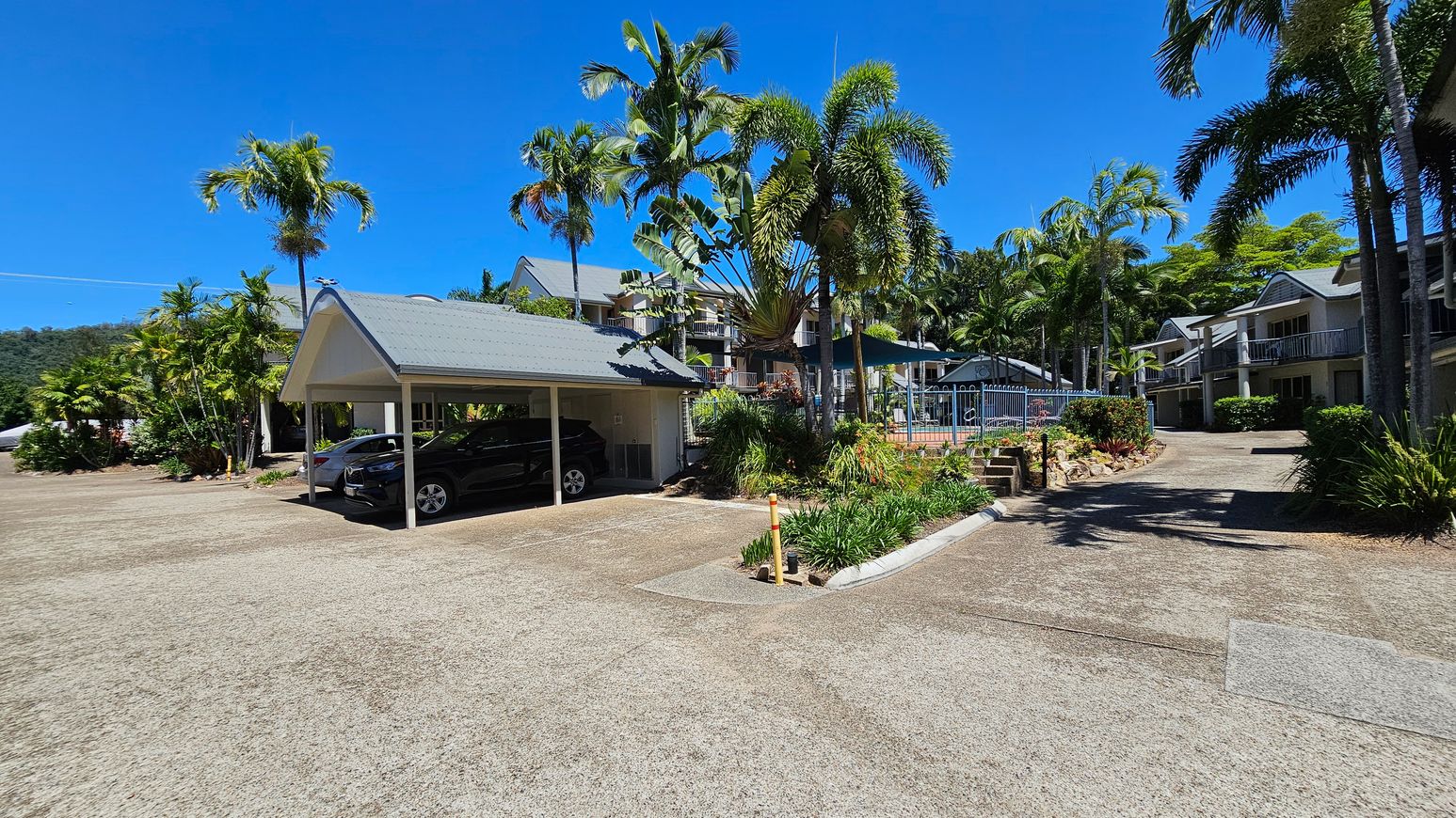 A carport on the left shelters two cars, surrounded by lush palm trees and greenery. In the centre, a pathway leads to a fenced pool area with more tropical plants. On the right, there are two-storey buildings with balconies, also surrounded by palm trees and shrubs. The ground is a mix of concrete and patches of grass. A carport on the left shelters two cars, surrounded by lush palm trees and greenery. In the centre, a pathway leads to a fenced pool area with more tropical plants. On the right, there are two-storey buildings with balconies, also surrounded by palm trees and shrubs. The ground is a mix of concrete and patches of grass.