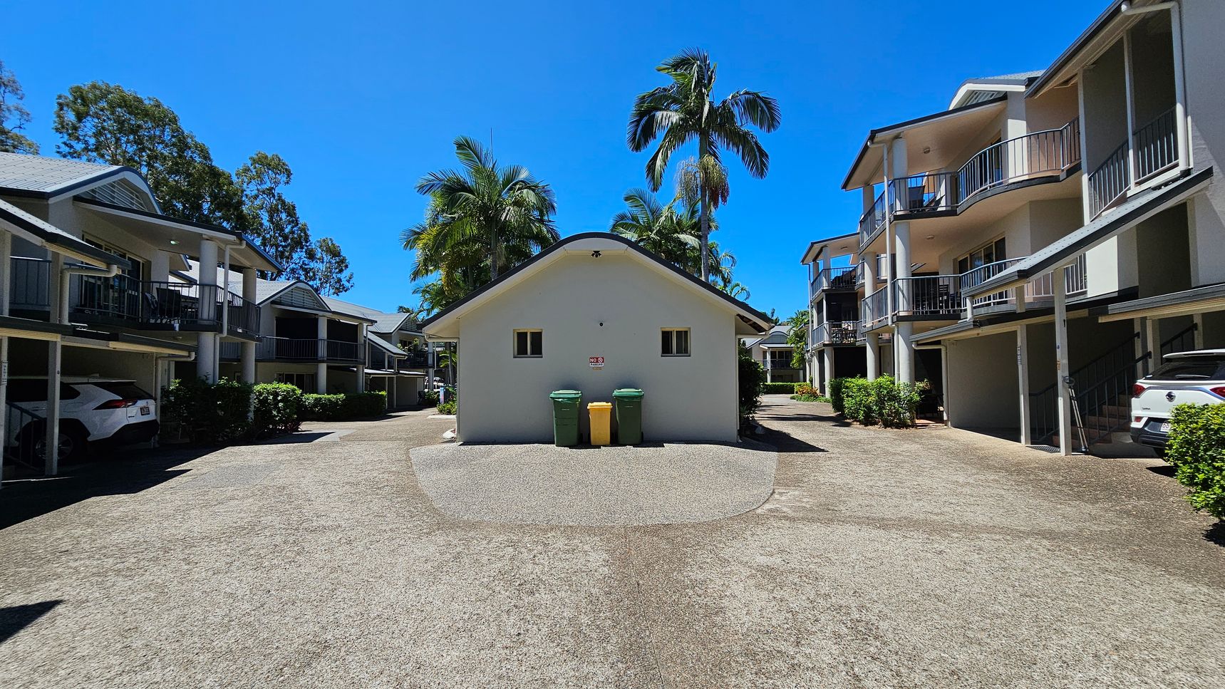 A courtyard with a central building featuring two small windows and three bins in front. On either side, there are two-storey residential buildings with balconies and carports. Cars are parked under the carports. Palm trees and other greenery are visible around the area. The ground is a mix of concrete and gravel. A courtyard with a central building featuring two small windows and three bins in front. On either side, there are two-storey residential buildings with balconies and carports. Cars are parked under the carports. Palm trees and other greenery are visible around the area. The ground is a mix of concrete and gravel.