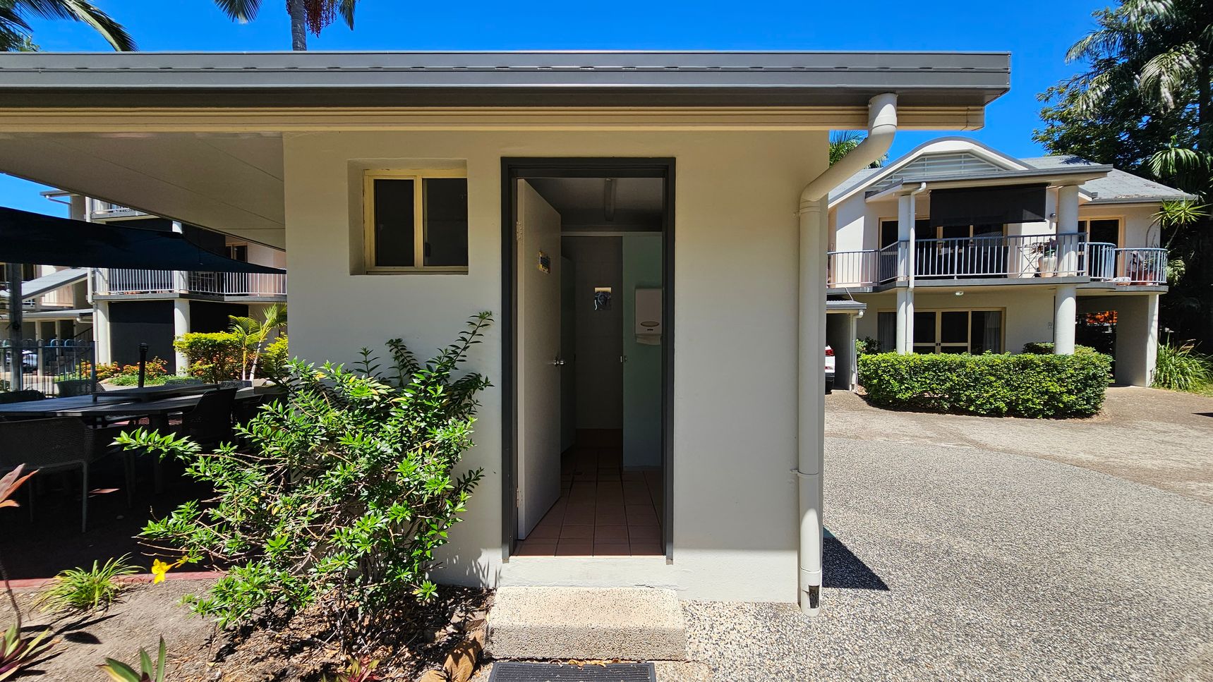 A small, light-coloured building with a narrow entrance in the centre. To the left, there are green shrubs and a shaded outdoor seating area with tables and chairs. On the right, a larger two-storey building with balconies and a hedge in front. The ground is a mix of concrete and gravel, and there are palm trees in the background. A small, light-coloured building with a narrow entrance in the centre. To the left, there are green shrubs and a shaded outdoor seating area with tables and chairs. On the right, a larger two-storey building with balconies and a hedge in front. The ground is a mix of concrete and gravel, and there are palm trees in the background.