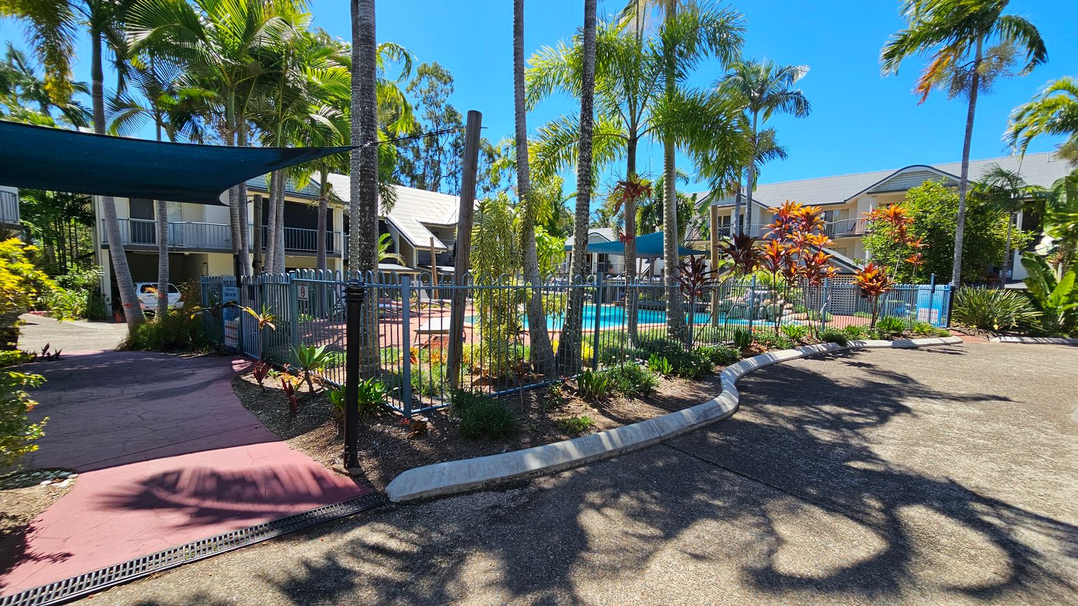 A tropical outdoor setting with a swimming pool surrounded by a metal fence in the centre. Tall palm trees and various plants are scattered around the area. On the left, there is a shaded pathway leading to a building with a parked car. On the right, more plants and a curved concrete barrier are visible. In the background, there are two-storey buildings with balconies. The ground is paved, and the sky is clear. A tropical outdoor setting with a swimming pool surrounded by a metal fence in the centre. Tall palm trees and various plants are scattered around the area. On the left, there is a shaded pathway leading to a building with a parked car. On the right, more plants and a curved concrete barrier are visible. In the background, there are two-storey buildings with balconies. The ground is paved, and the sky is clear.