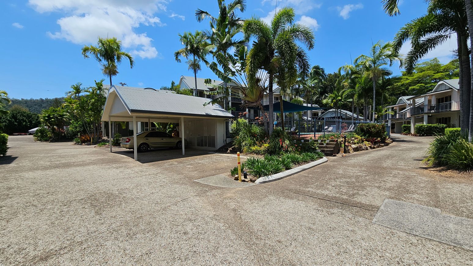 A paved area with a carport on the left, sheltering a parked car. Palm trees and lush greenery surround the area. In the centre, a pathway leads to a fenced pool area with a shade structure. On the right, there are two-storey buildings with balconies, surrounded by more palm trees and shrubs. The sky is clear with a few clouds. A paved area with a carport on the left, sheltering a parked car. Palm trees and lush greenery surround the area. In the centre, a pathway leads to a fenced pool area with a shade structure. On the right, there are two-storey buildings with balconies, surrounded by more palm trees and shrubs. The sky is clear with a few clouds.