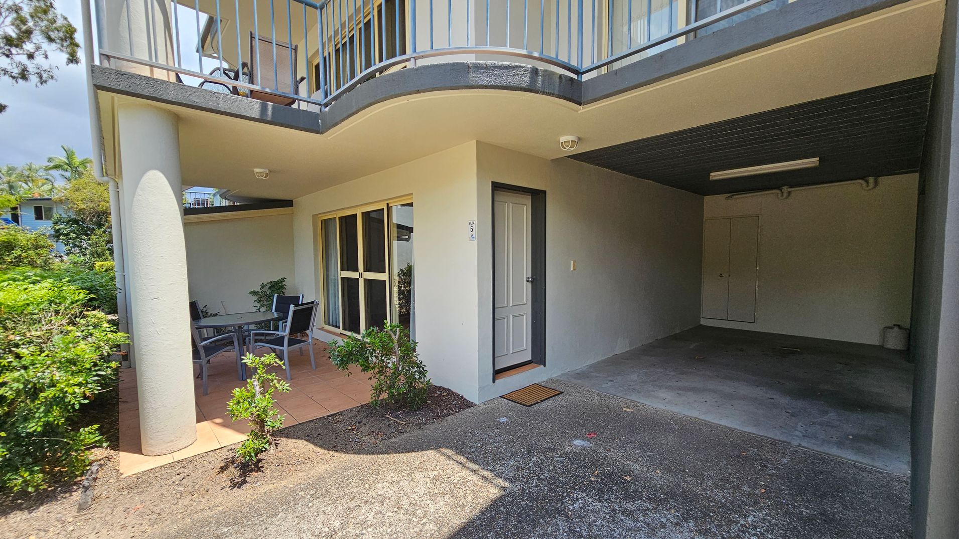 A ground-level outdoor area with a covered patio on the left, featuring a table and chairs. The patio is bordered by a low wall and greenery. A sliding glass door and a regular door are visible on the wall. On the right, there's a covered carport with a concrete floor and a small storage door at the back. The ceiling is flat with a light fixture. Above, a balcony with a metal railing is visible. The ground is a mix of concrete and tiled surfaces. A ground-level outdoor area with a covered patio on the left, featuring a table and chairs. The patio is bordered by a low wall and greenery. A sliding glass door and a regular door are visible on the wall. On the right, there's a covered carport with a concrete floor and a small storage door at the back. The ceiling is flat with a light fixture. Above, a balcony with a metal railing is visible. The ground is a mix of concrete and tiled surfaces.