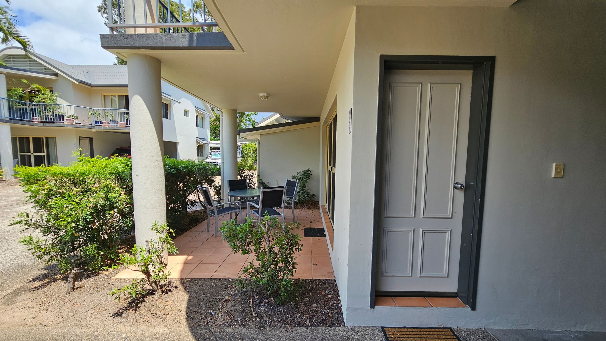 A covered patio area with a tiled floor and a round glass table surrounded by four chairs. To the right, there's a closed door with a doormat in front. The patio is bordered by a low hedge and a few small plants. In the background, there are two-storey buildings with balconies and potted plants. A driveway is visible on the left side. The ceiling of the patio is plain and has a light fixture. A covered patio area with a tiled floor and a round glass table surrounded by four chairs. To the right, there's a closed door with a doormat in front. The patio is bordered by a low hedge and a few small plants. In the background, there are two-storey buildings with balconies and potted plants. A driveway is visible on the left side. The ceiling of the patio is plain and has a light fixture.